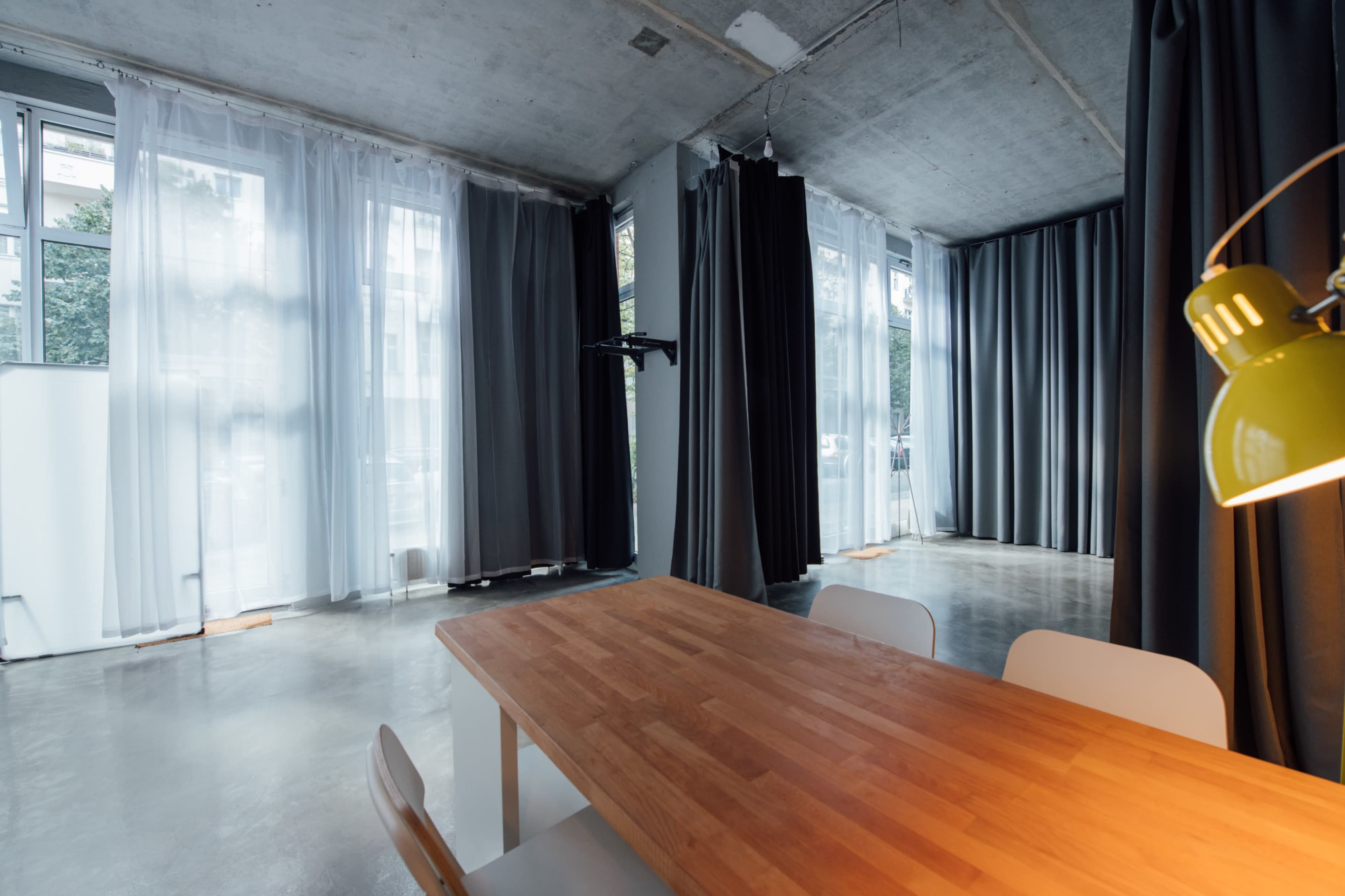 A spacious room with large windows features a wooden table surrounded by white chairs and both sheer and dark curtains covering the windows.