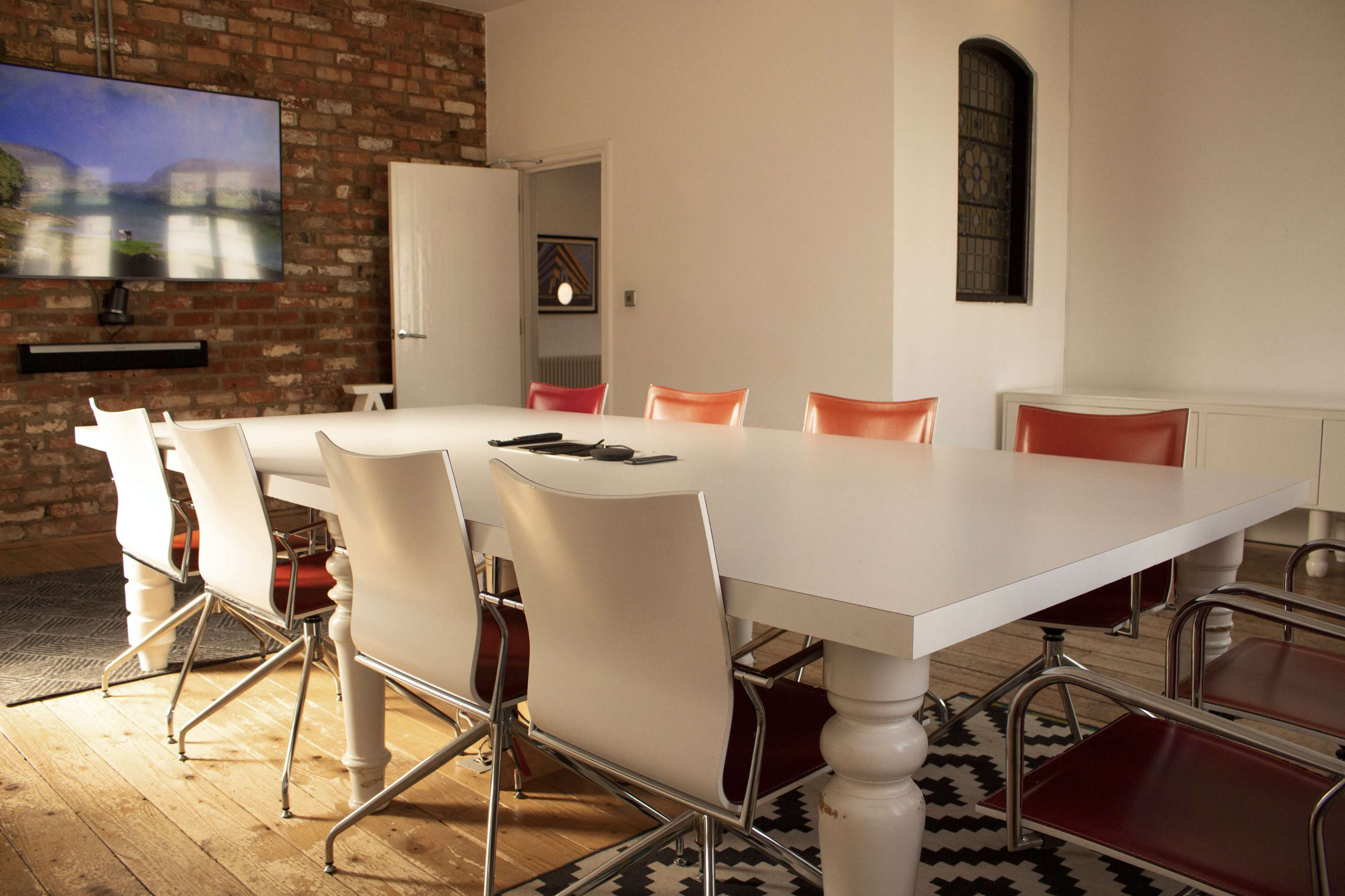 A large, white conference table surrounded by several chairs is set in a room with exposed brick walls and a television mounted on one wall.