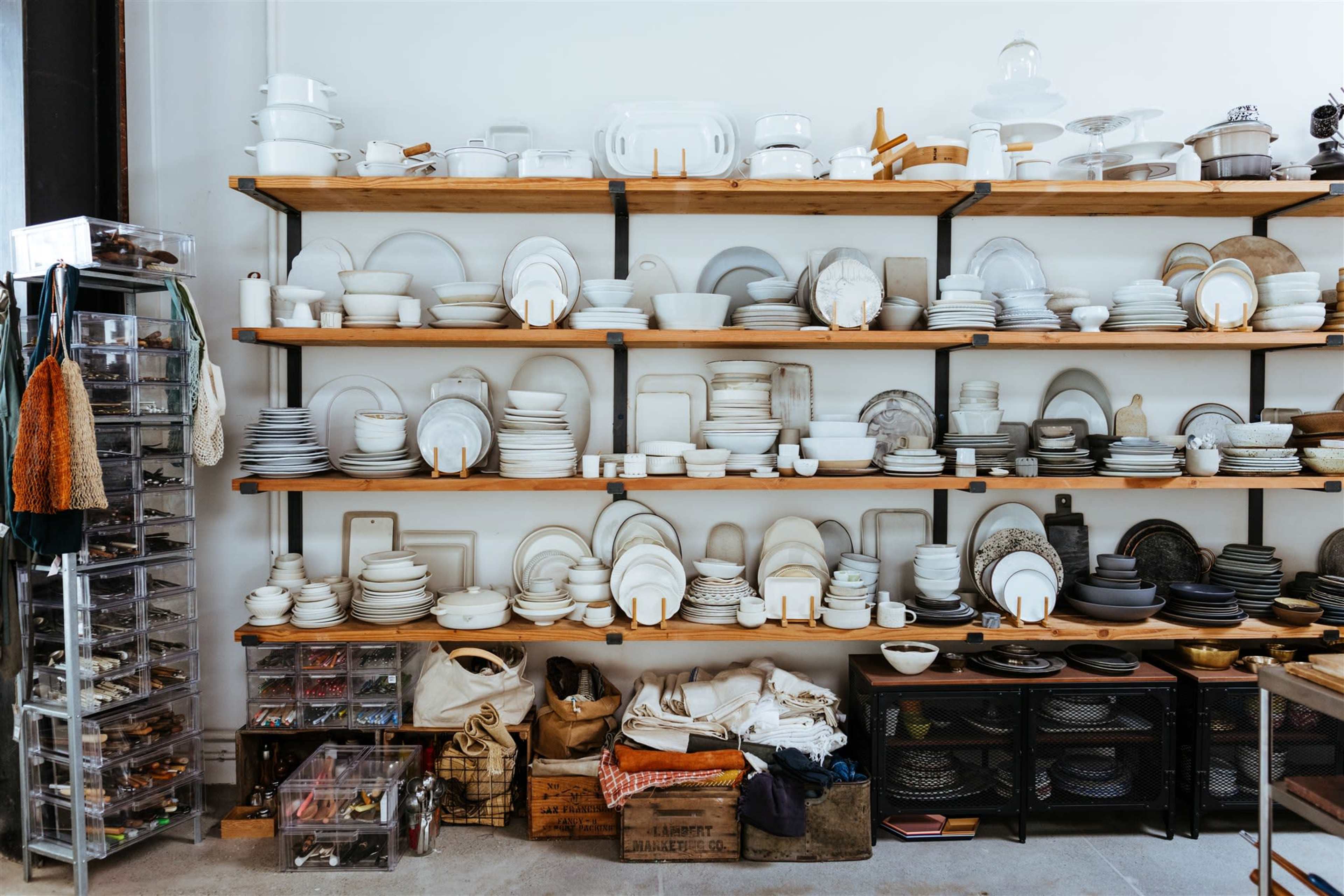 A cluttered display of various dishes and kitchenware arranged on wooden shelves, with storage bins and containers underneath.