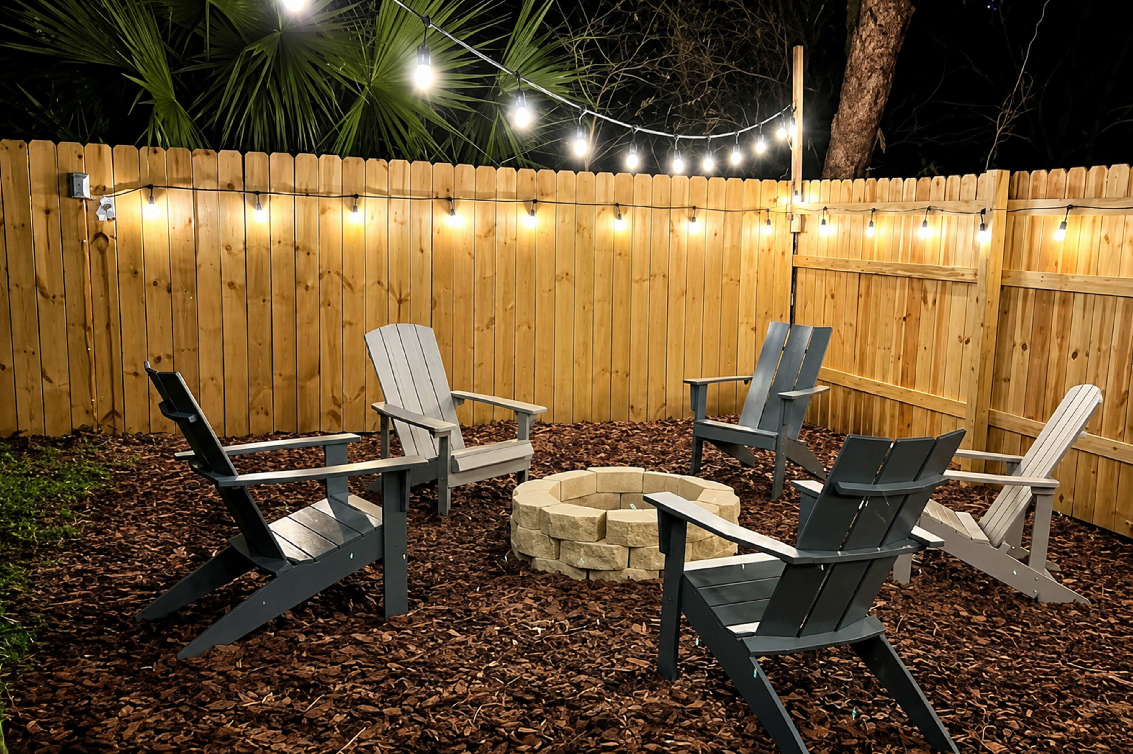 The image shows a cozy outdoor fire pit area with four chairs arranged around a stone circle, surrounded by mulch and a fenced wooden backdrop with string lights overhead.