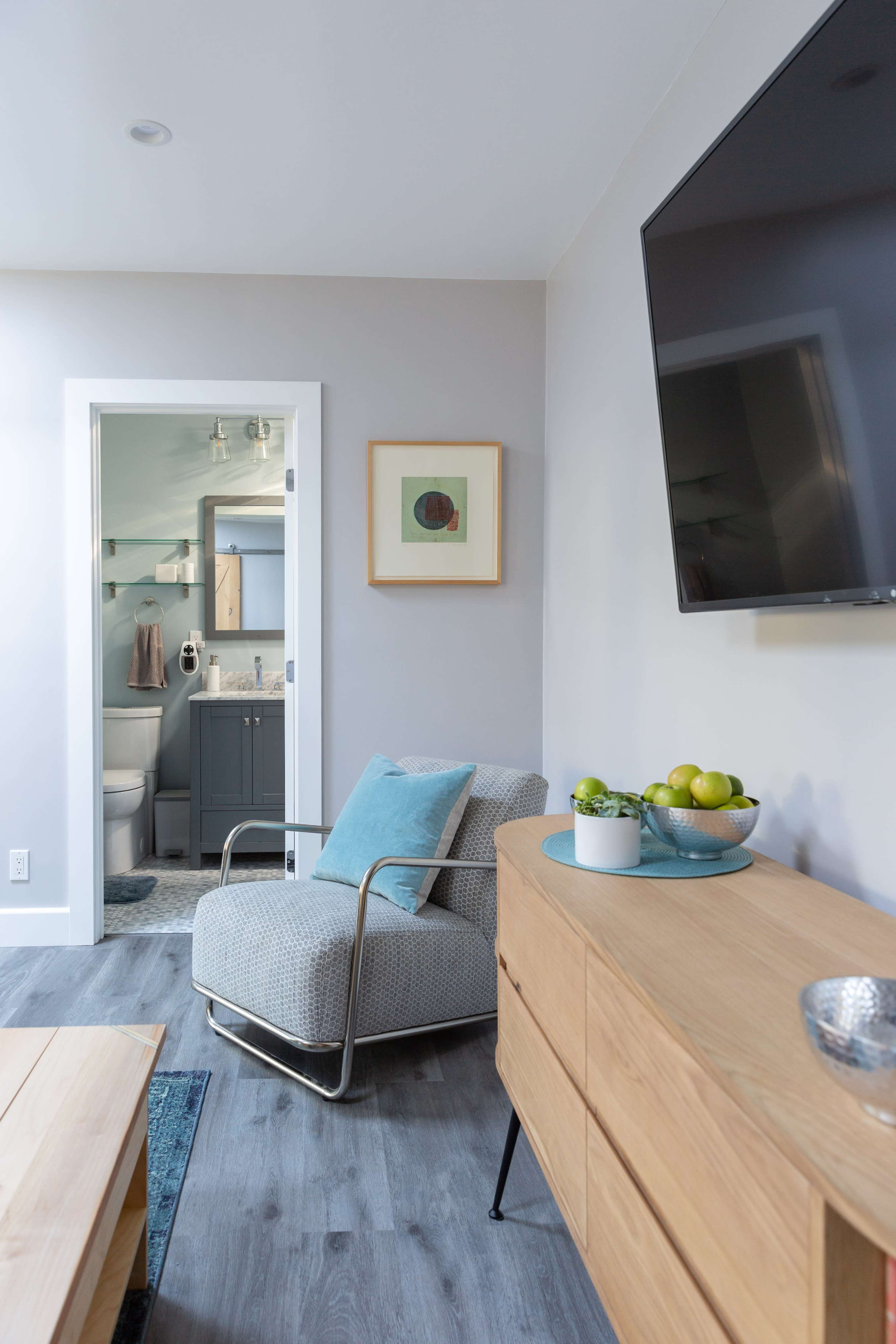 A modern living room features a chair and cabinet, with a view of a bathroom through an open door.