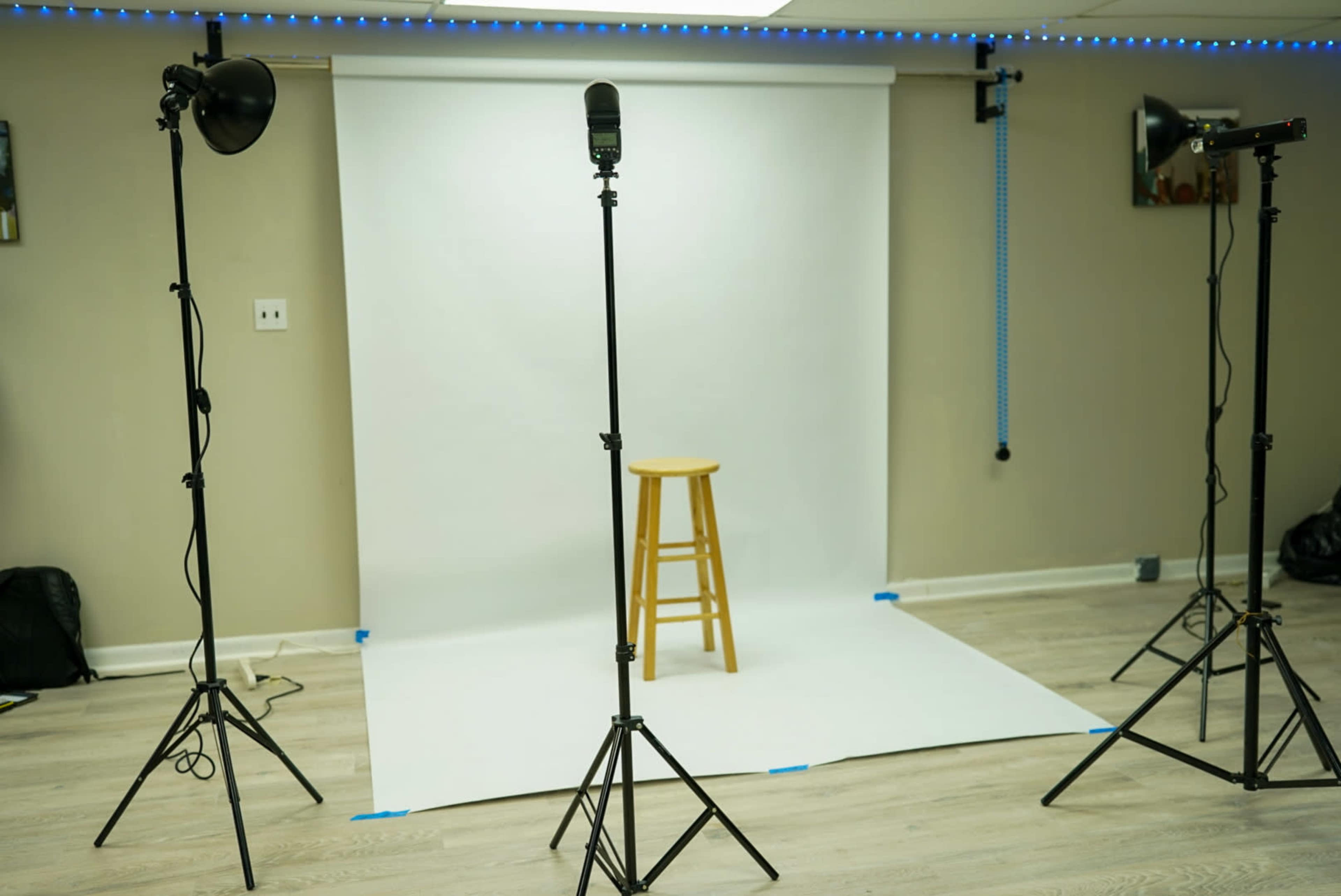 A photography studio setup features a white backdrop, a wooden stool in the center, and three lighting stands positioned around it.