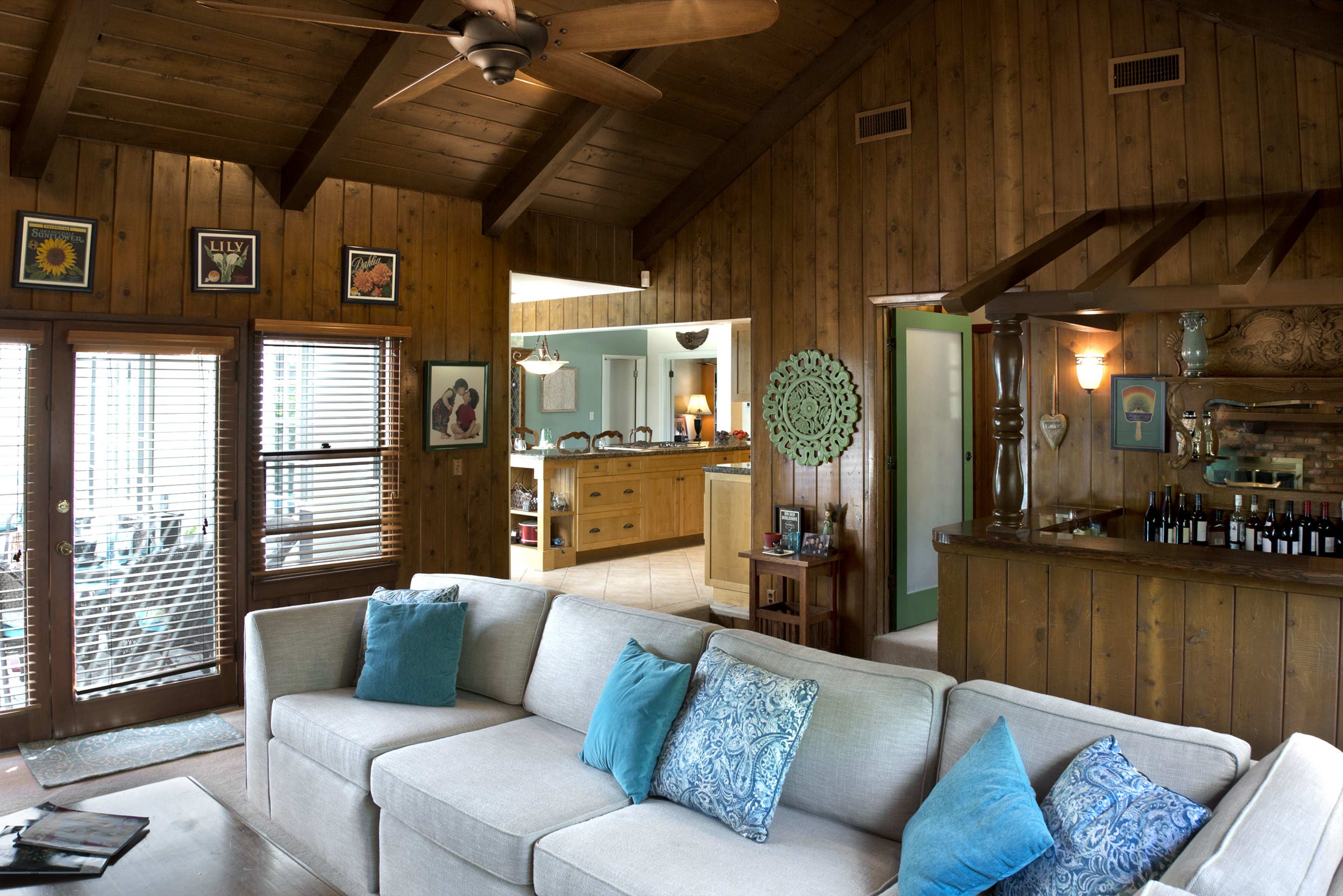 The image shows a living room with a beige couch adorned with blue pillows, surrounded by wood-paneled walls and an open view into a kitchen area.