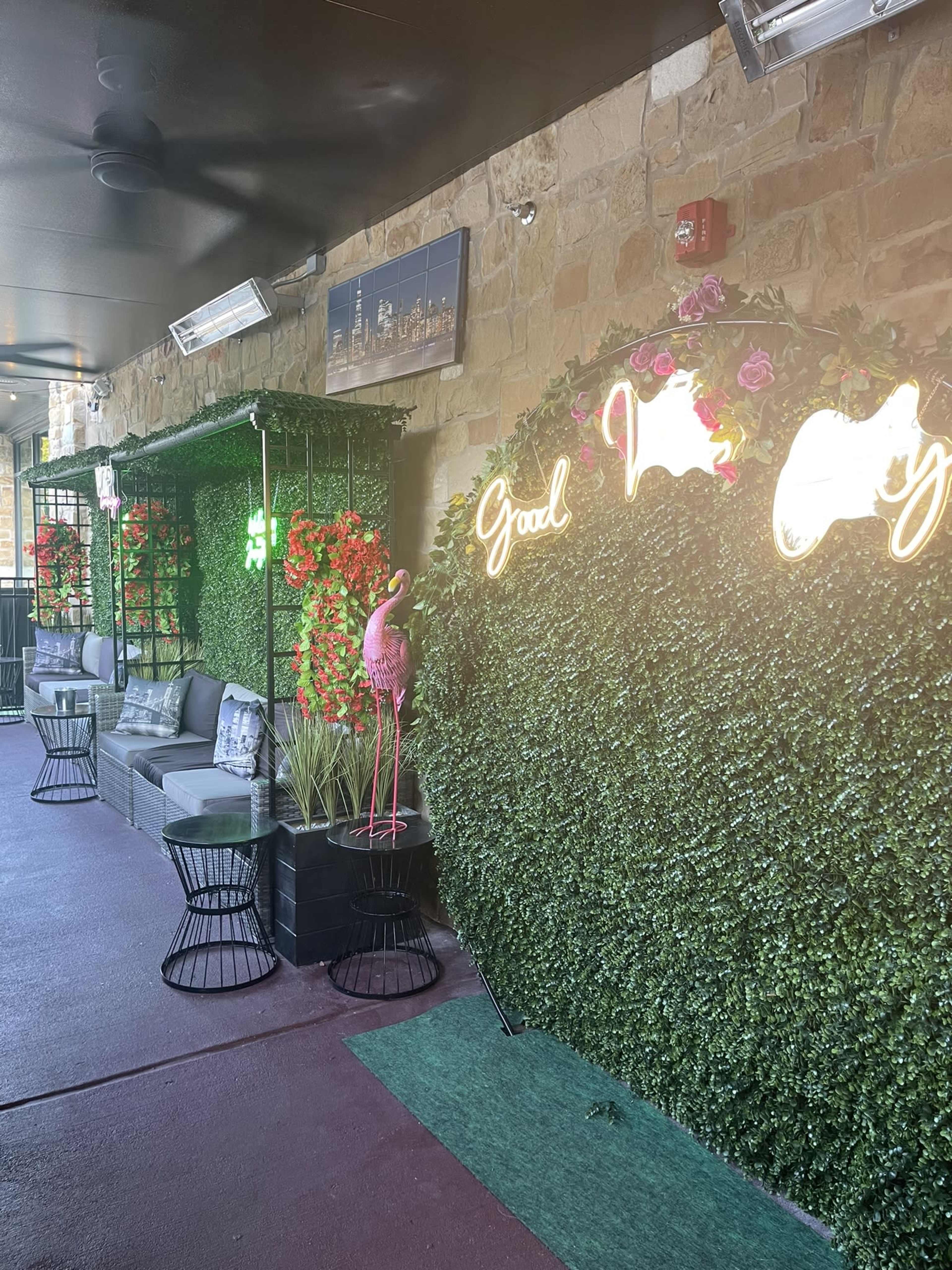 A lounge area decorated with green foliage, neon signs, and pink flamingo statues, featuring gray sofas and black wire tables.