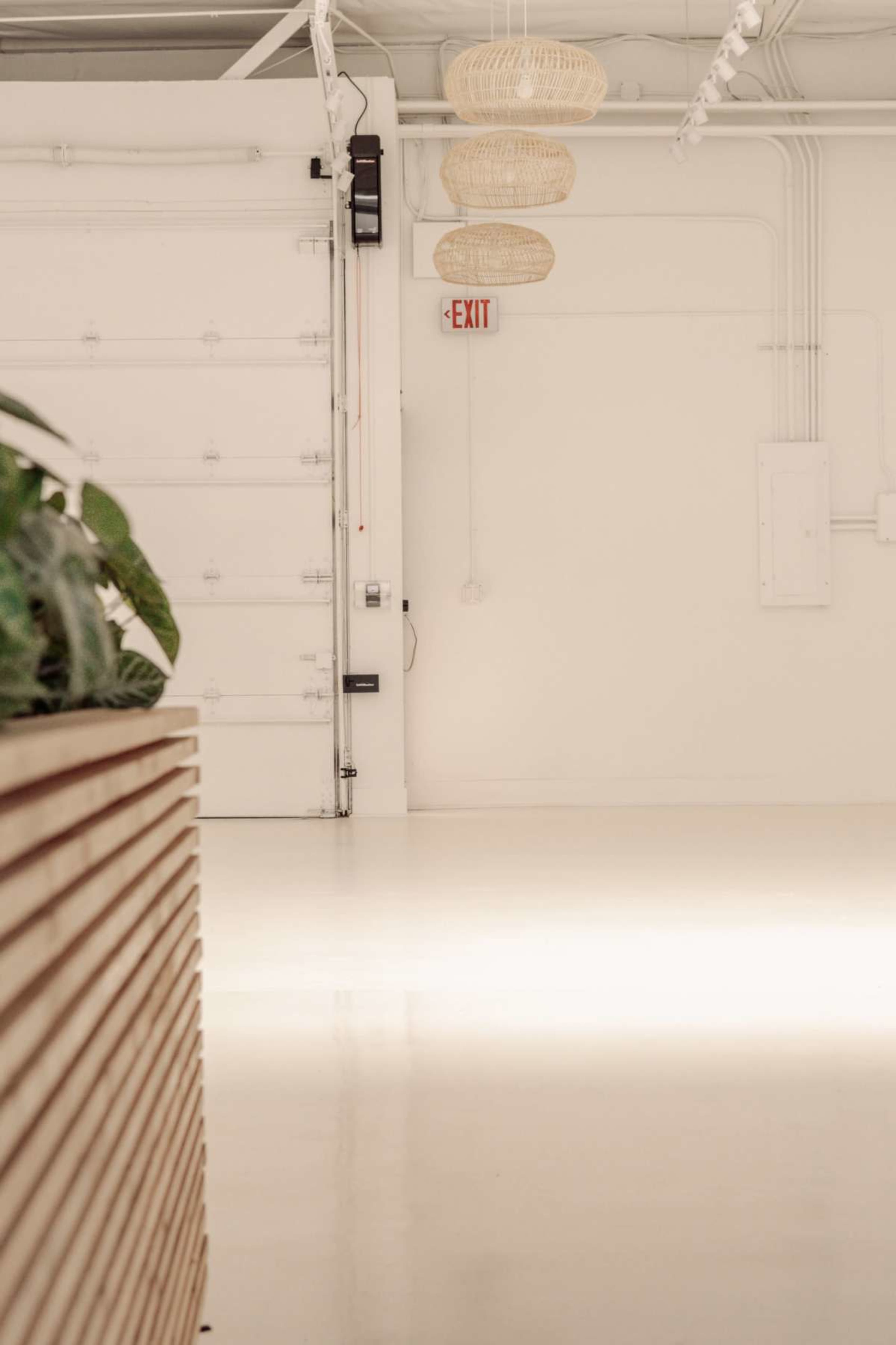 The image shows a spacious, minimalist interior with a large white wall, an exit sign, and three hanging light fixtures.