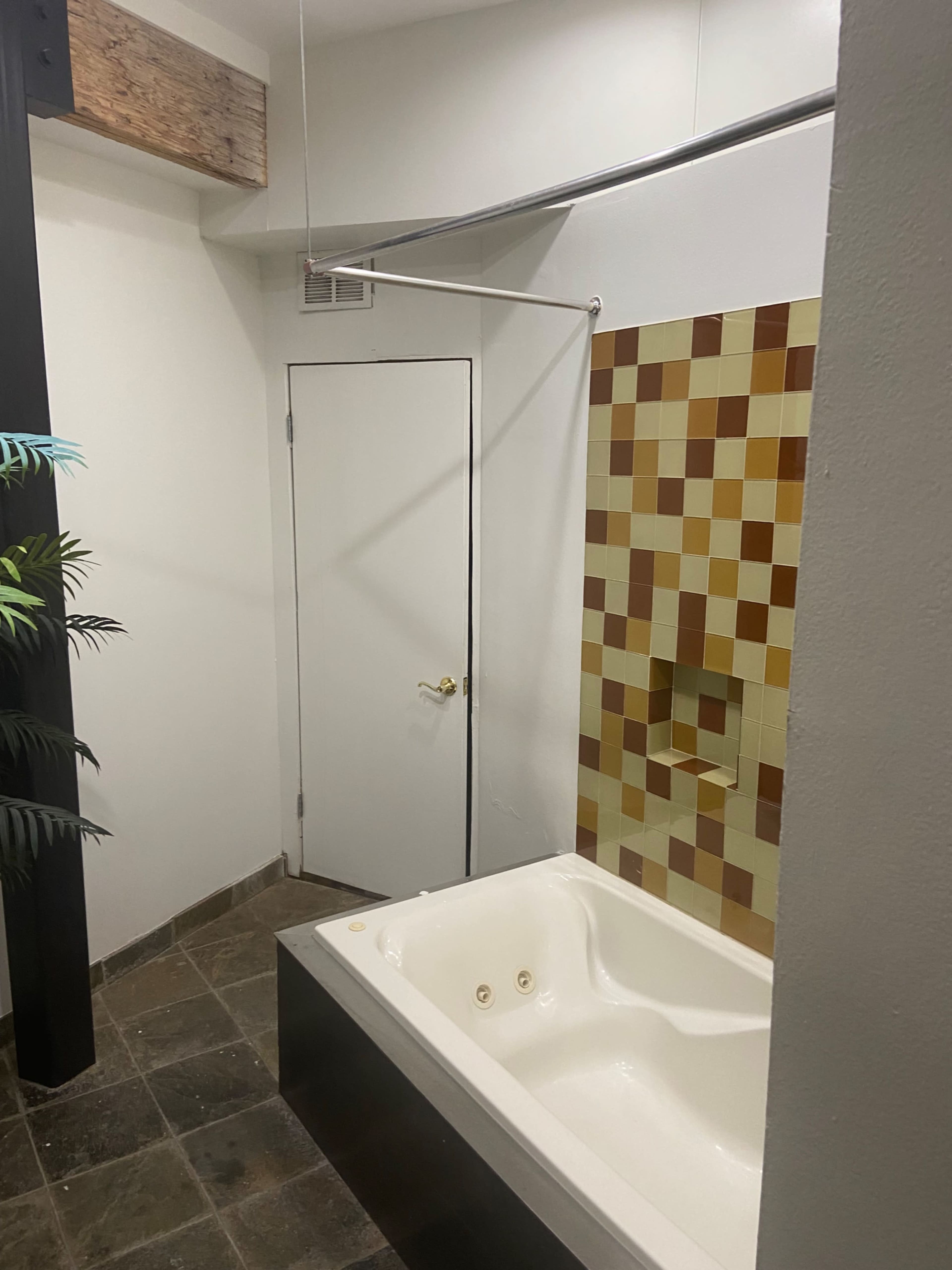 The image shows a bathroom featuring a white bathtub, a tiled wall in shades of brown and yellow, and a door leading to another area.