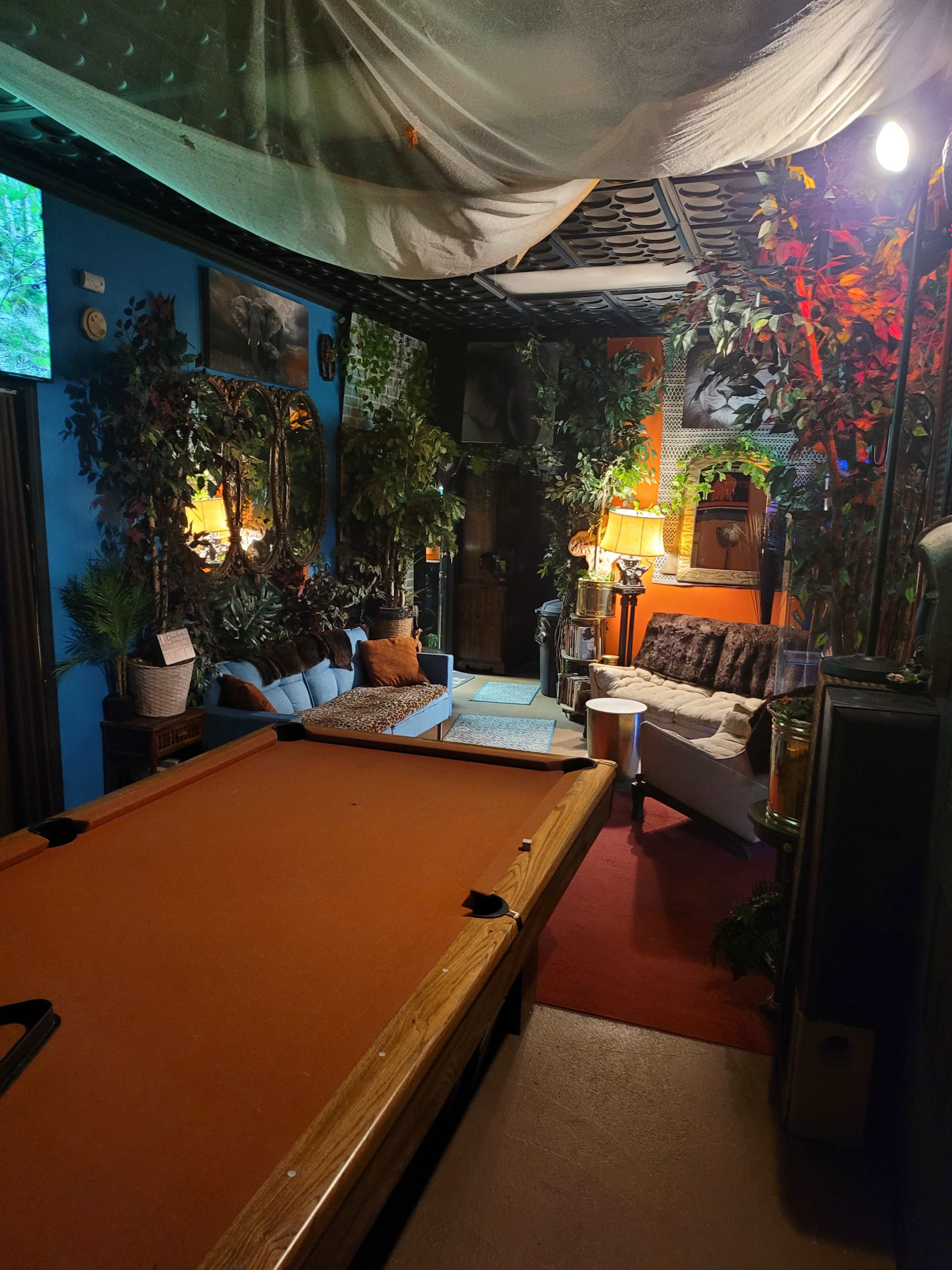 A cozy game room featuring a pool table, seating areas with plants, and warm lighting.