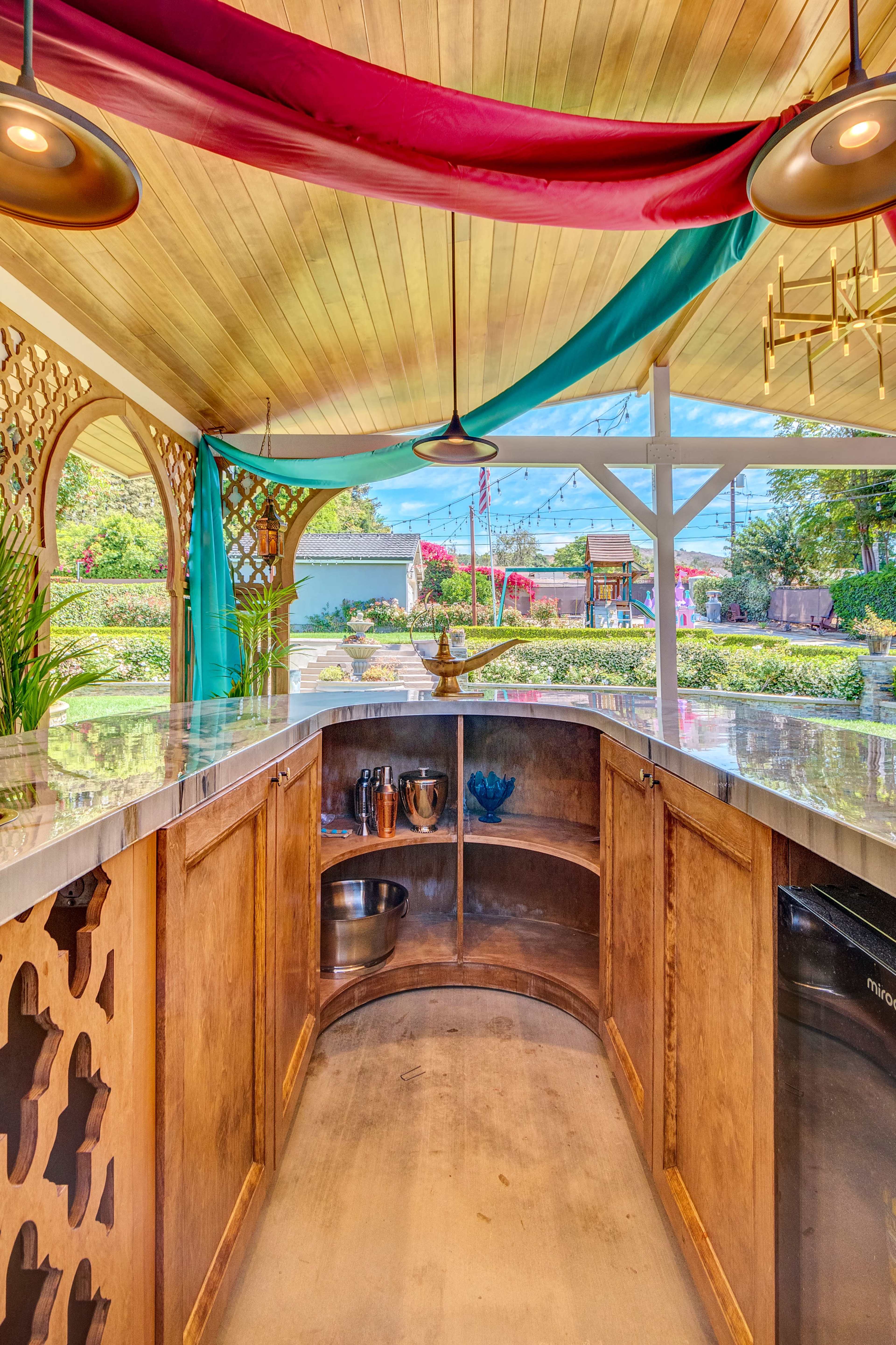 The image shows a well-designed outdoor bar with a curved wooden counter, decorative wall features, and colorful drapes overhead.