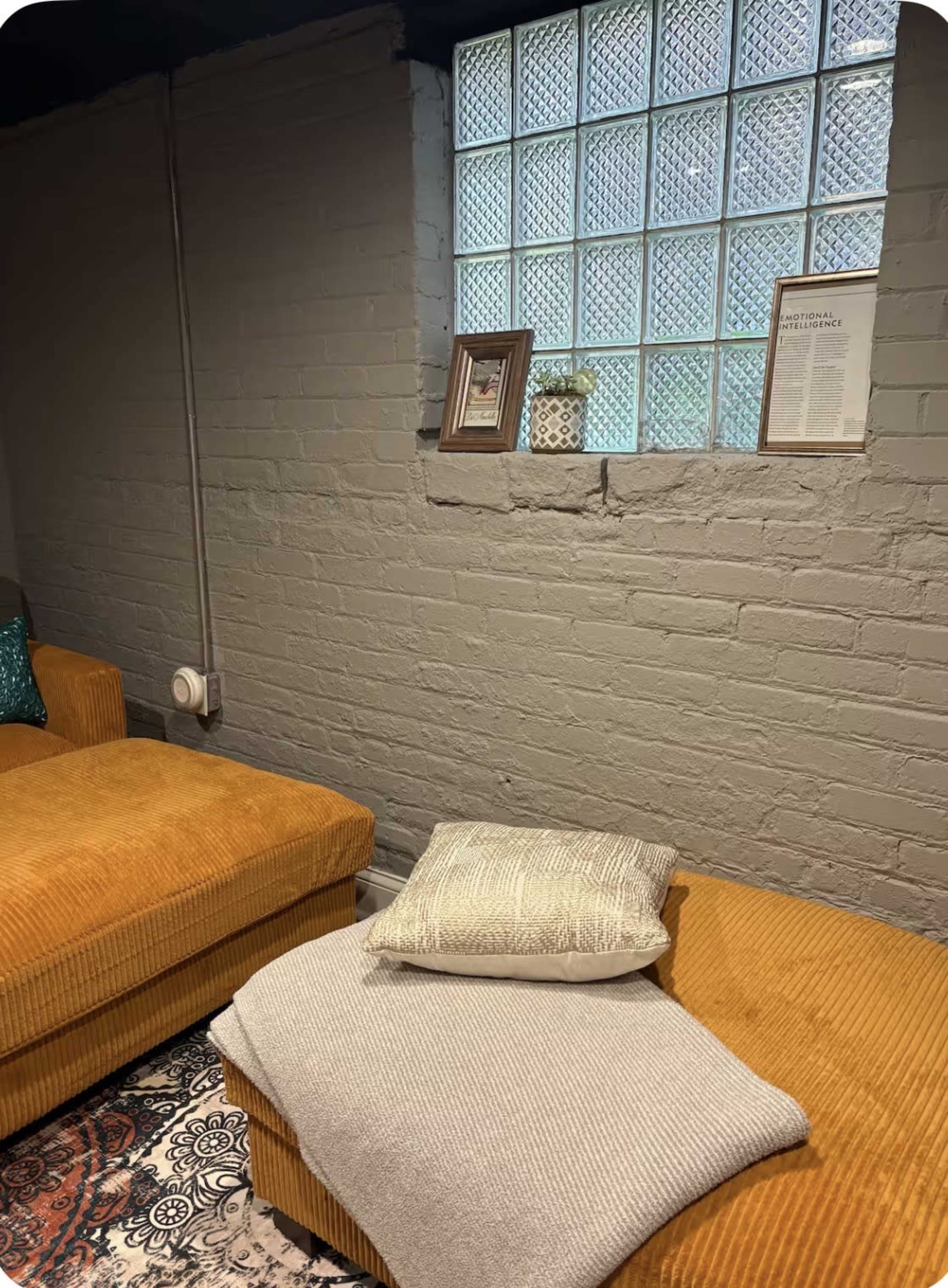 A cozy seating area features a large yellow couch with a blanket and a decorative pillow, alongside a window adorned with glass blocks and framed pictures.