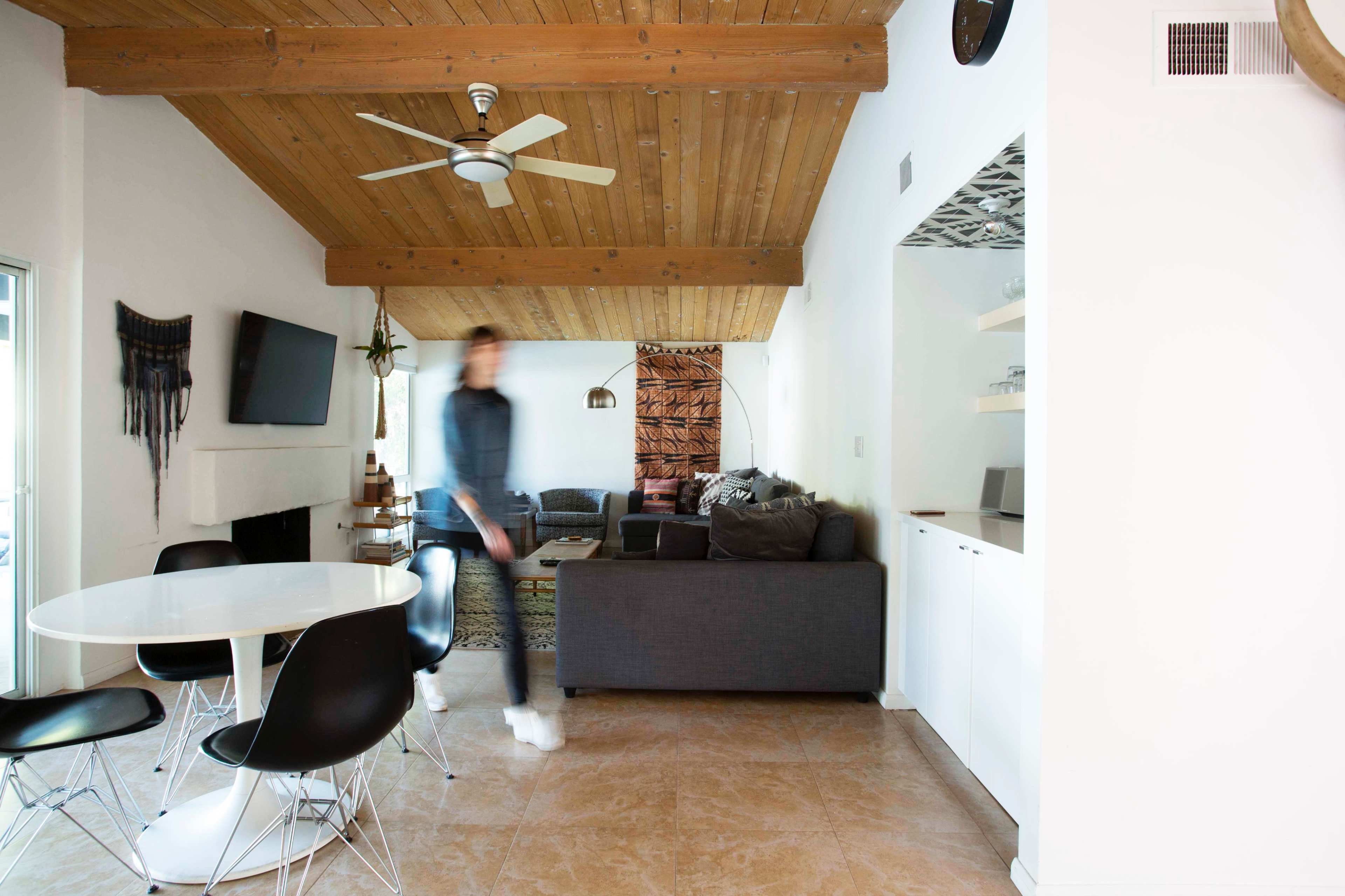 A person walks through a living space featuring a ceiling with wooden beams, a television mounted on the wall, and a dining table surrounded by black chairs.