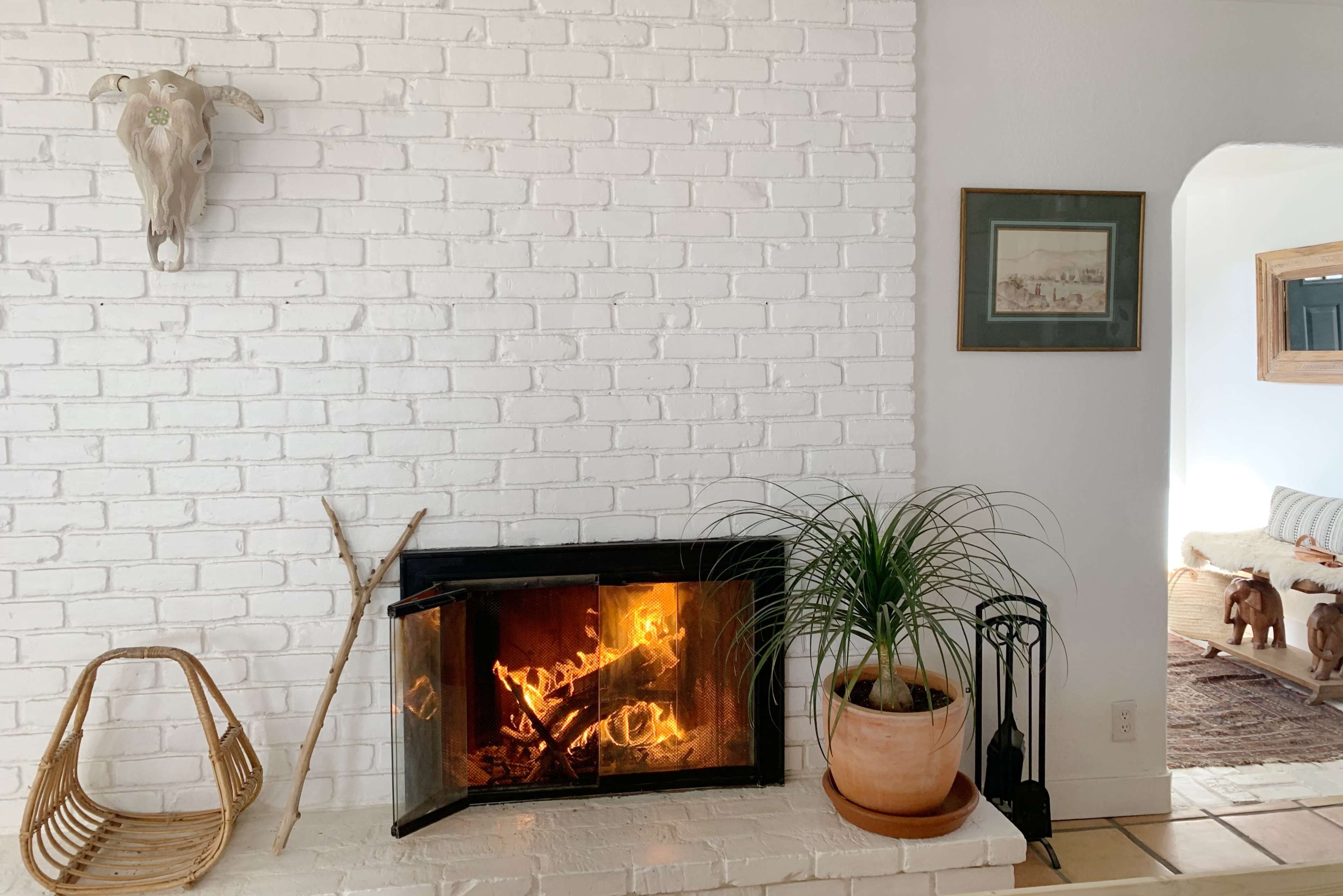 A white-brick fireplace with a lit fire and a potted plant sits beside a woven chair and an archway leading to another room.