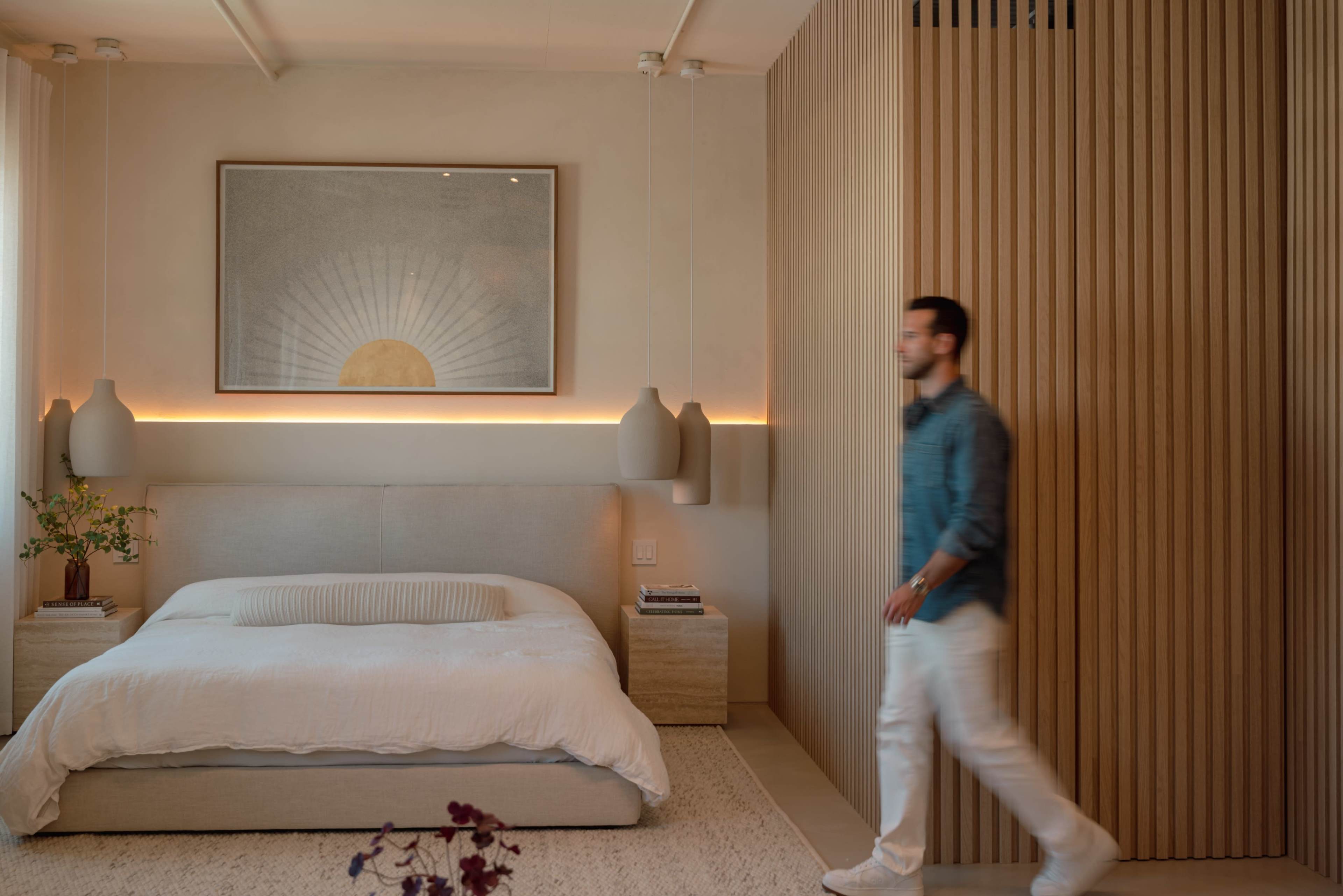 A man walks through a softly lit bedroom featuring a bed with a white duvet, wooden accent walls, and a large art piece depicting a sun.