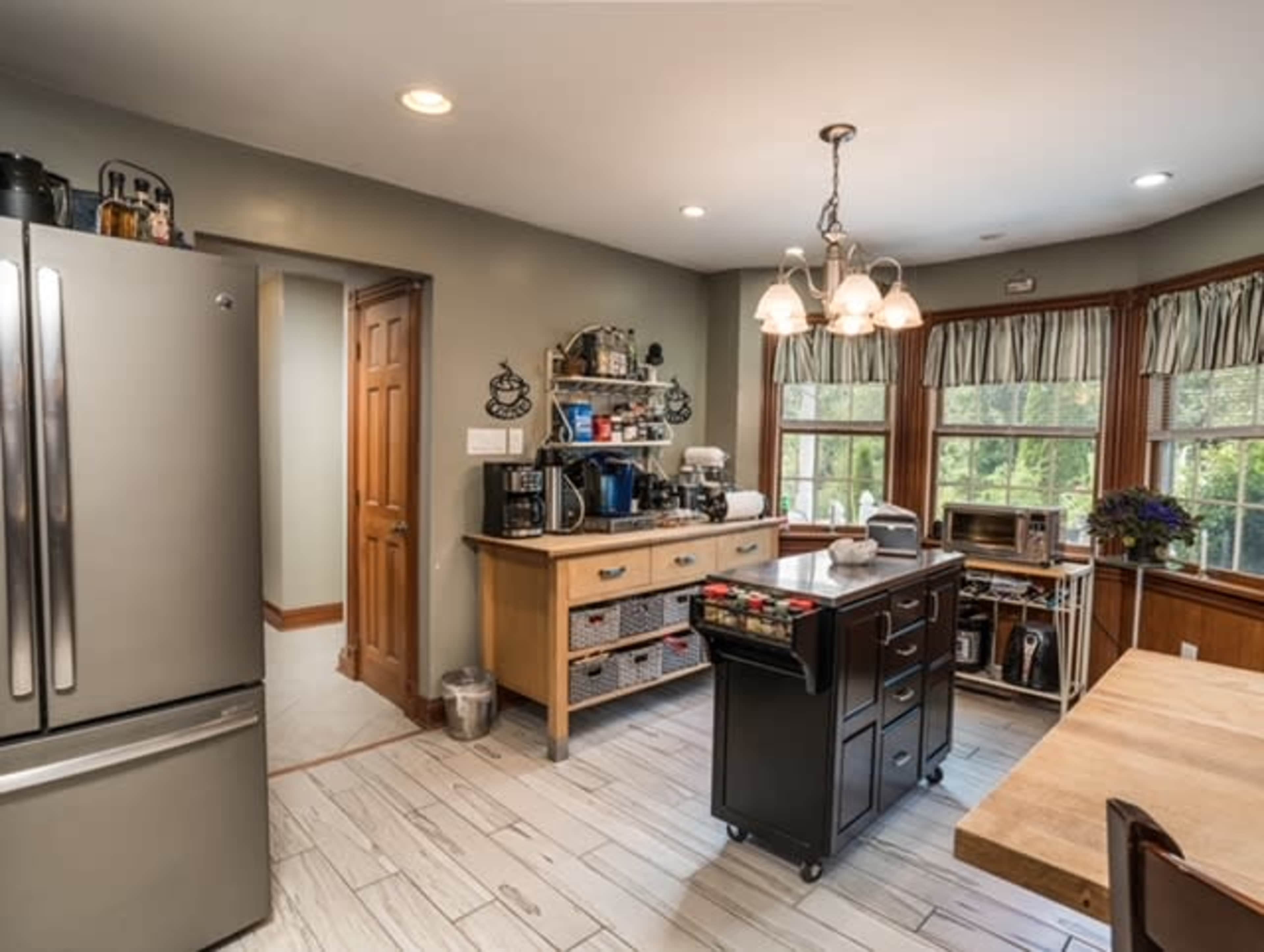 A kitchen features a stainless steel refrigerator, a wooden island with storage, coffee equipment, and large windows illuminating the space.