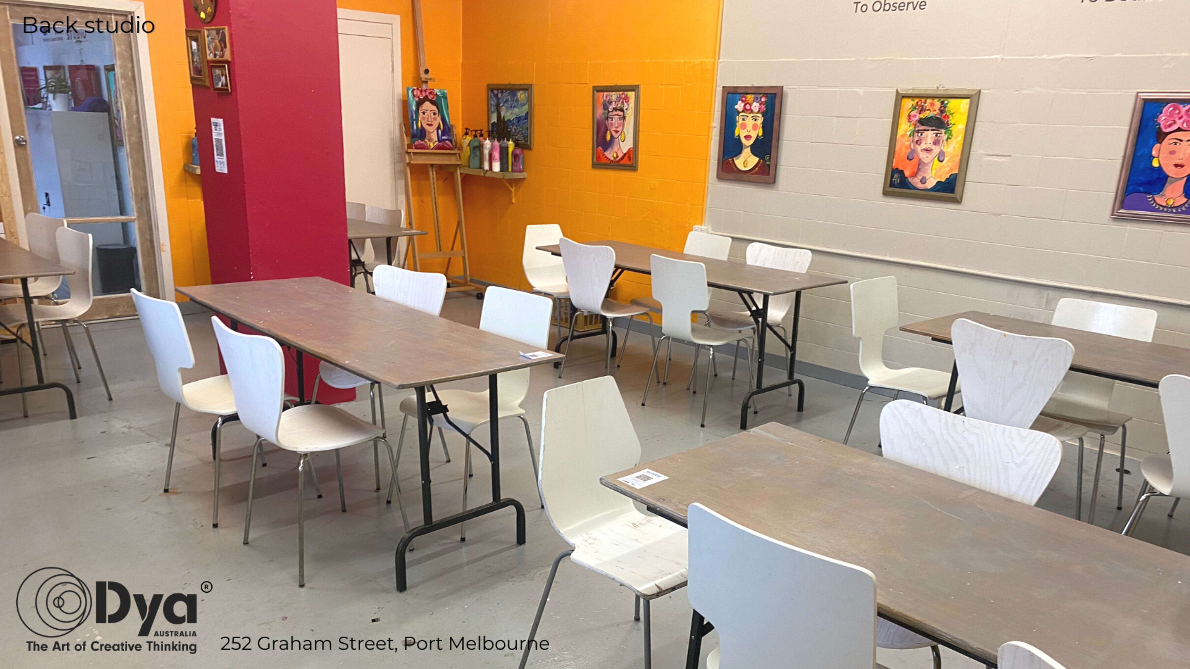 An art studio with several tables and chairs arranged around a brightly colored wall featuring framed artworks.