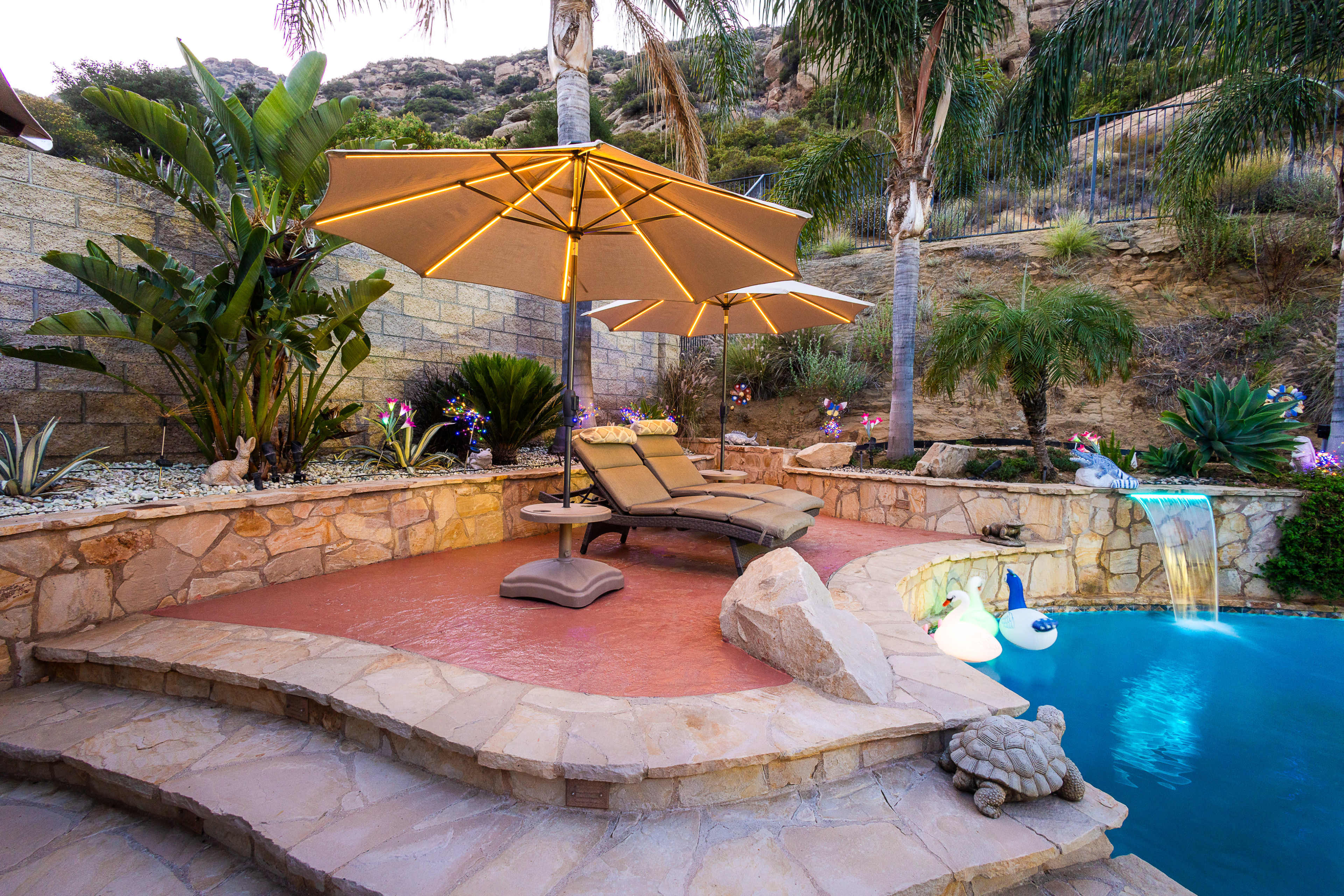 A poolside area features lounge chairs under two large umbrellas, surrounded by tropical plants and illuminated by soft lighting.