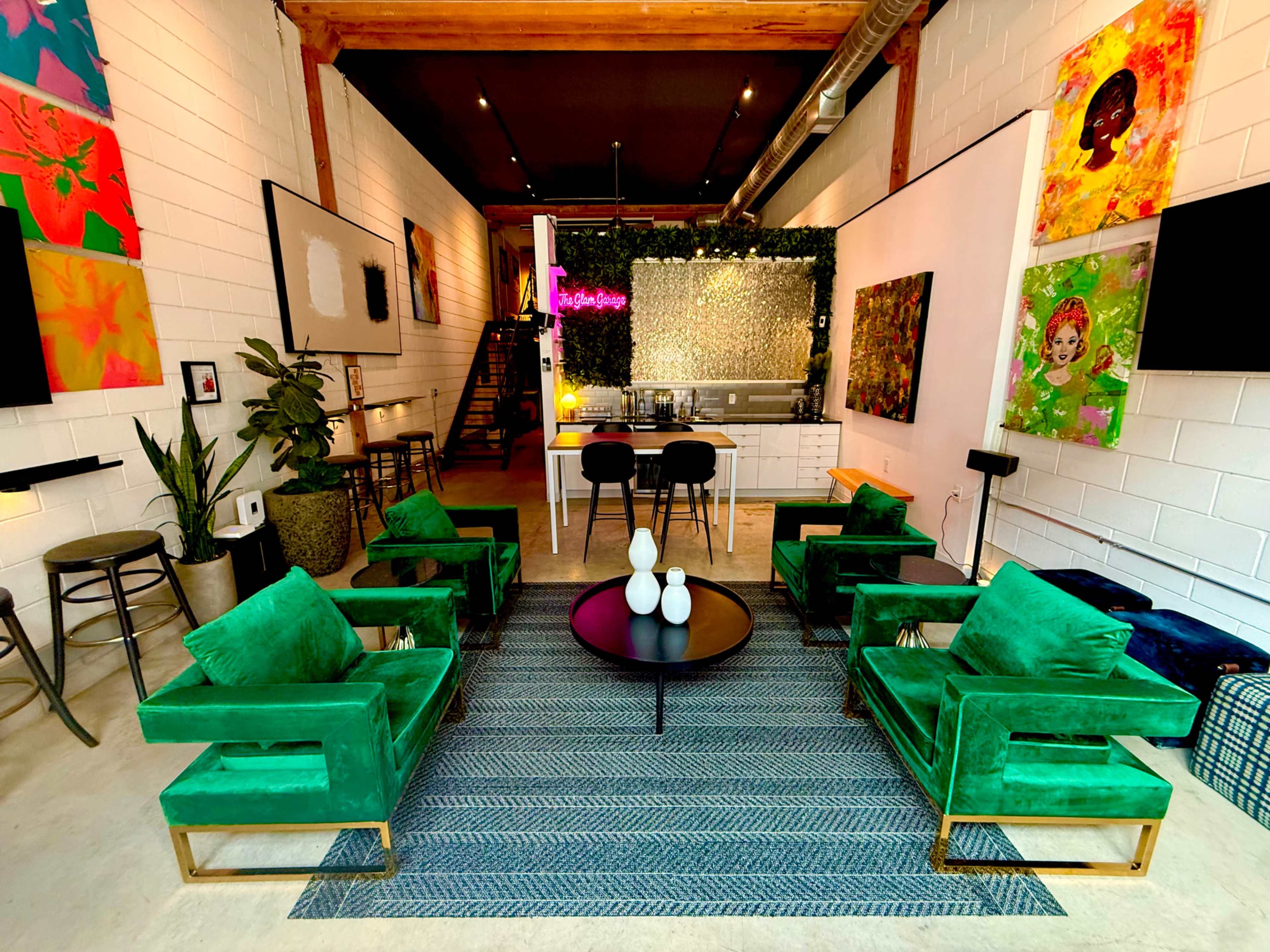 The image depicts a modern interior space with green velvet chairs arranged around a round coffee table, featuring colorful artwork on the walls and a kitchen area in the background.
