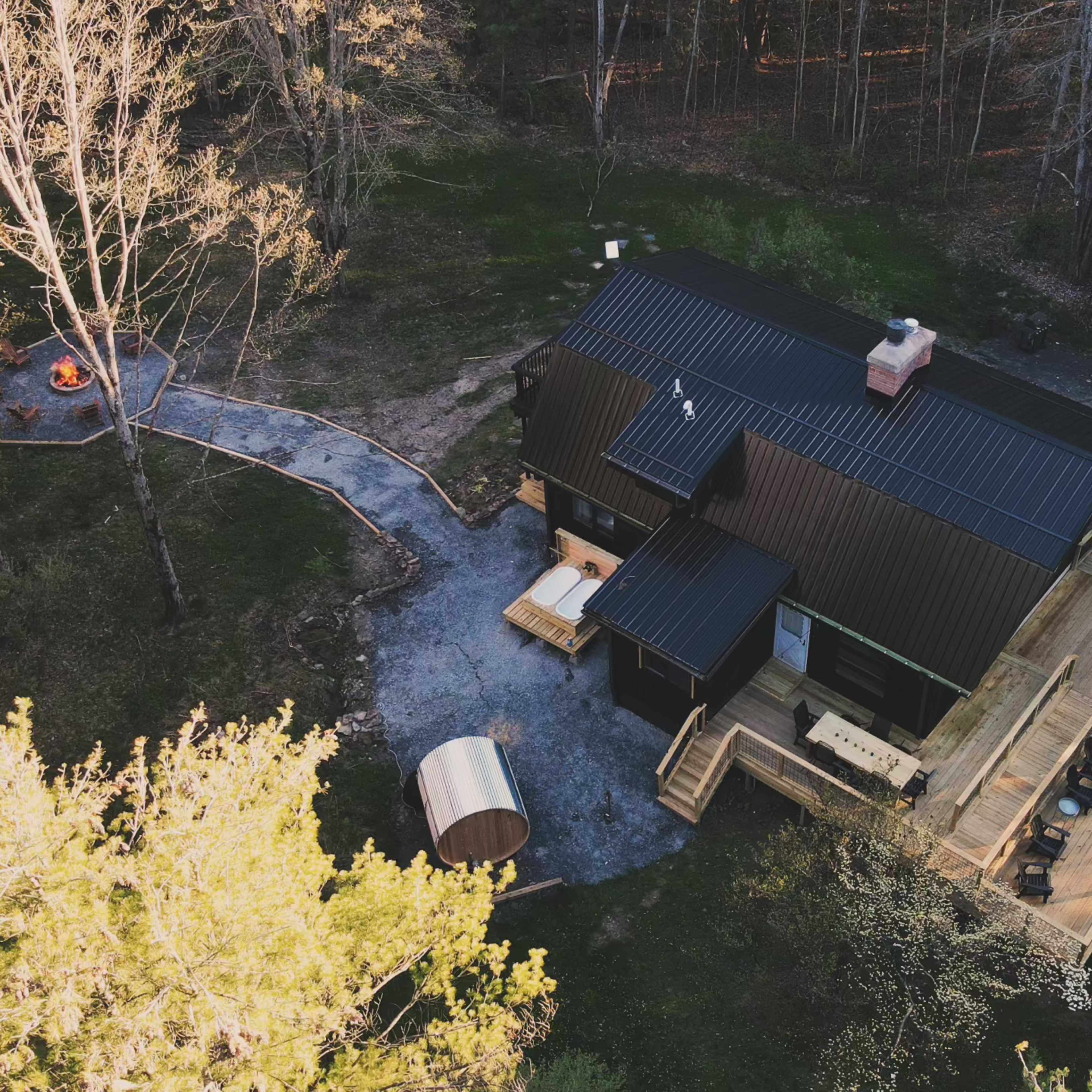 A modern black house with a wooden deck, a hot tub, and a fire pit surrounded by trees.