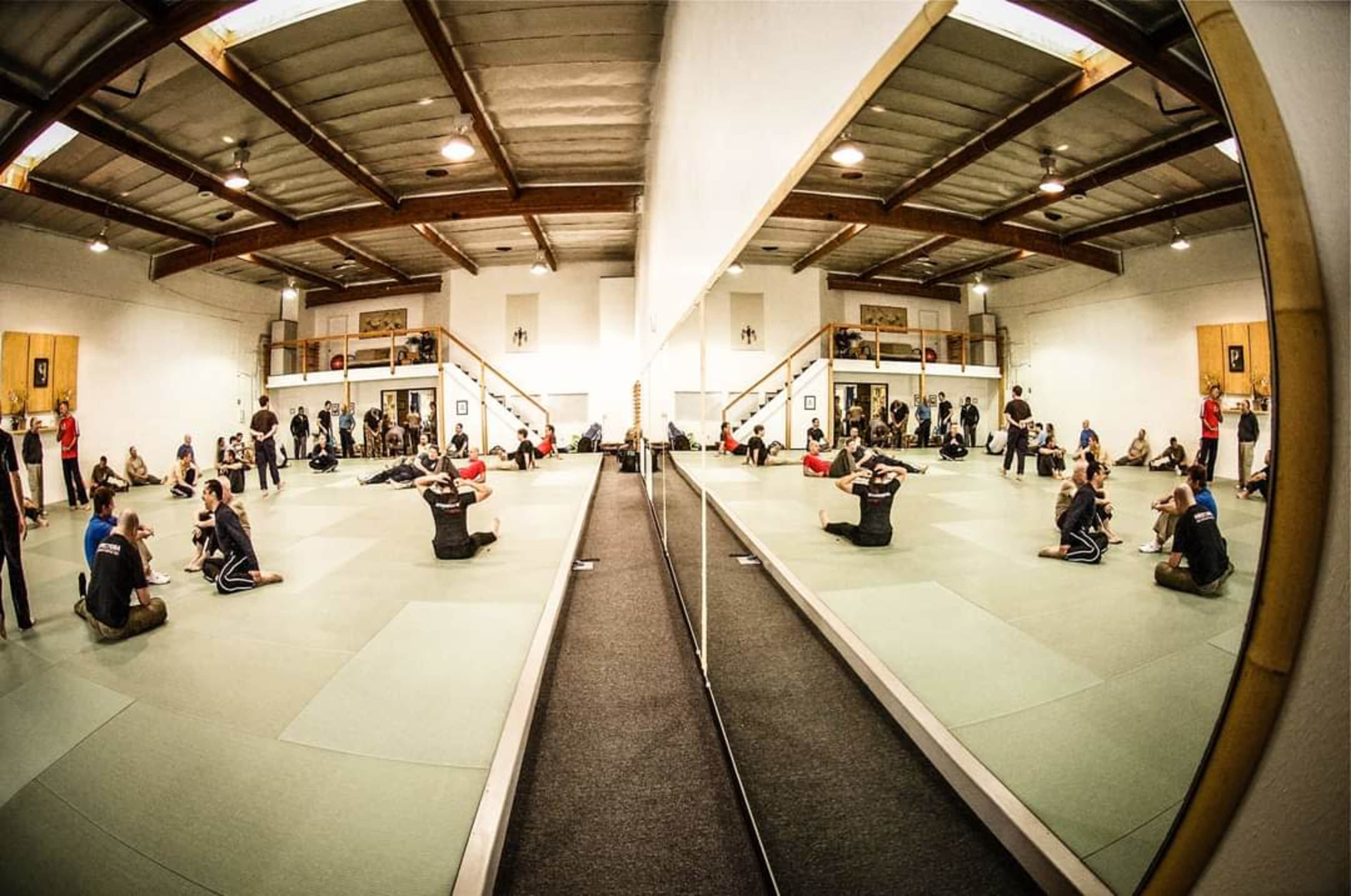 A martial arts training facility with multiple practitioners engaged in various training exercises on green mats, reflected in large wall mirrors.