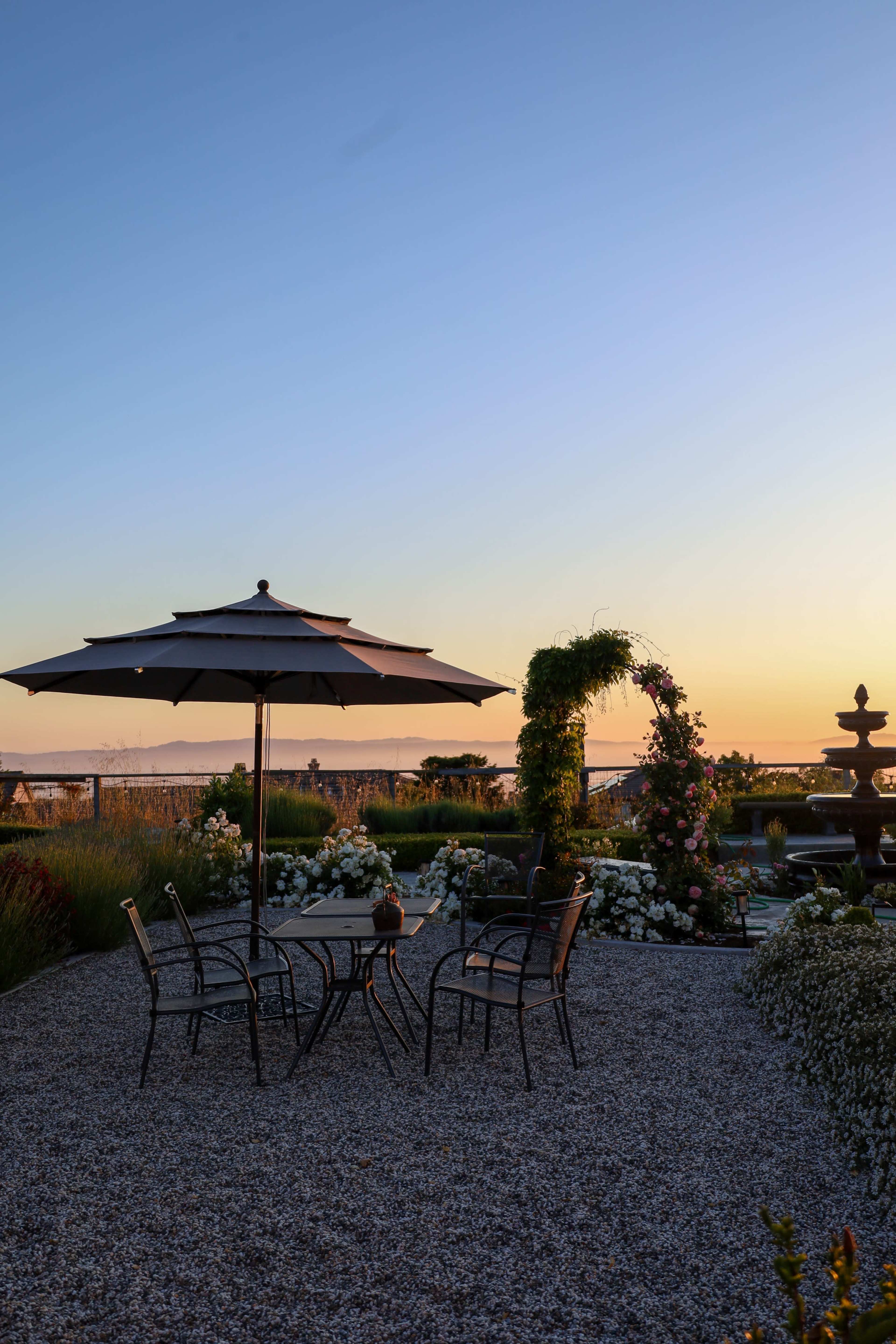 A patio with a table and chairs is shaded by an umbrella, overlooking a garden with flowers and a fountain against a sunset sky.