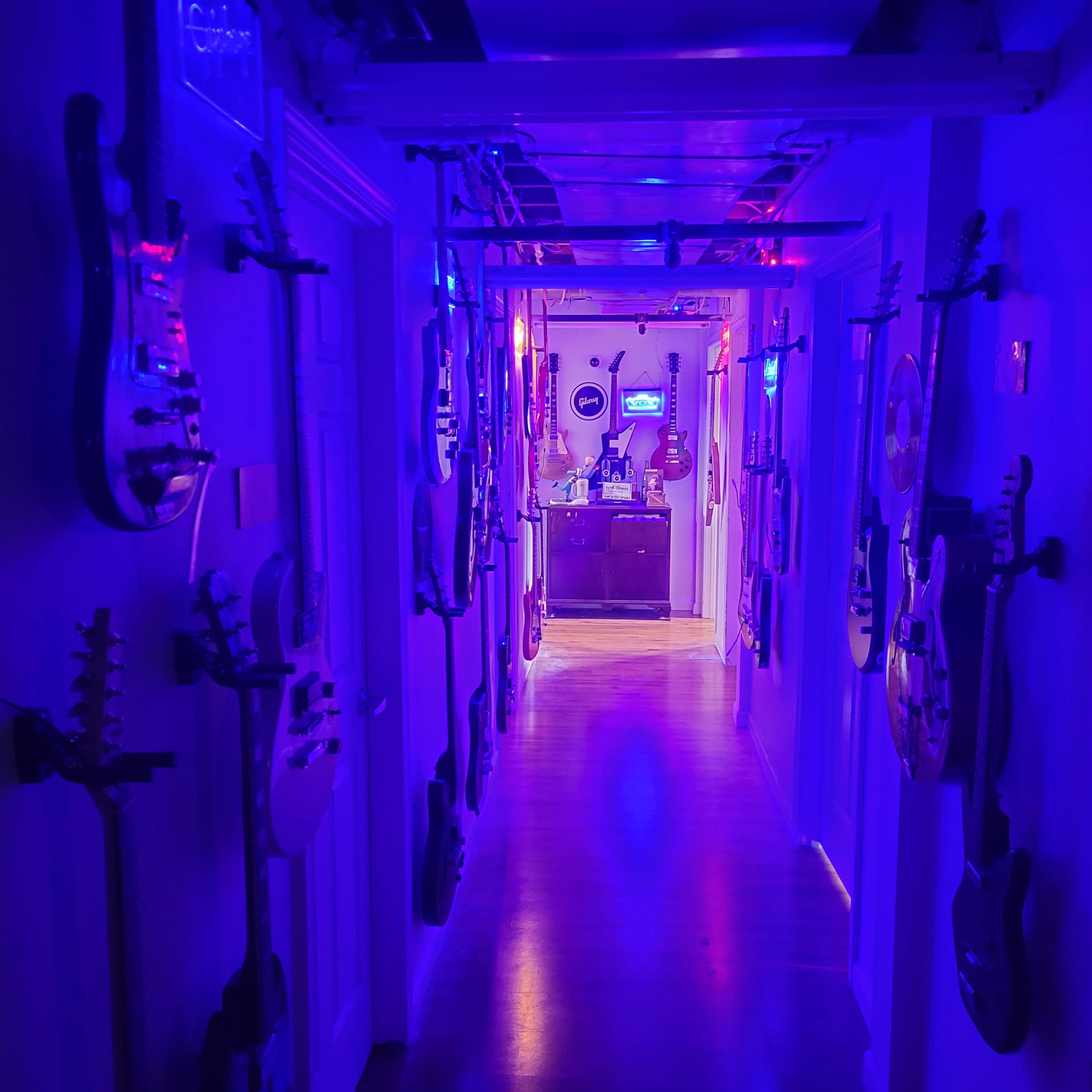 A narrow hallway lined with electric guitars on both sides leads to a room illuminated with blue and purple lights.