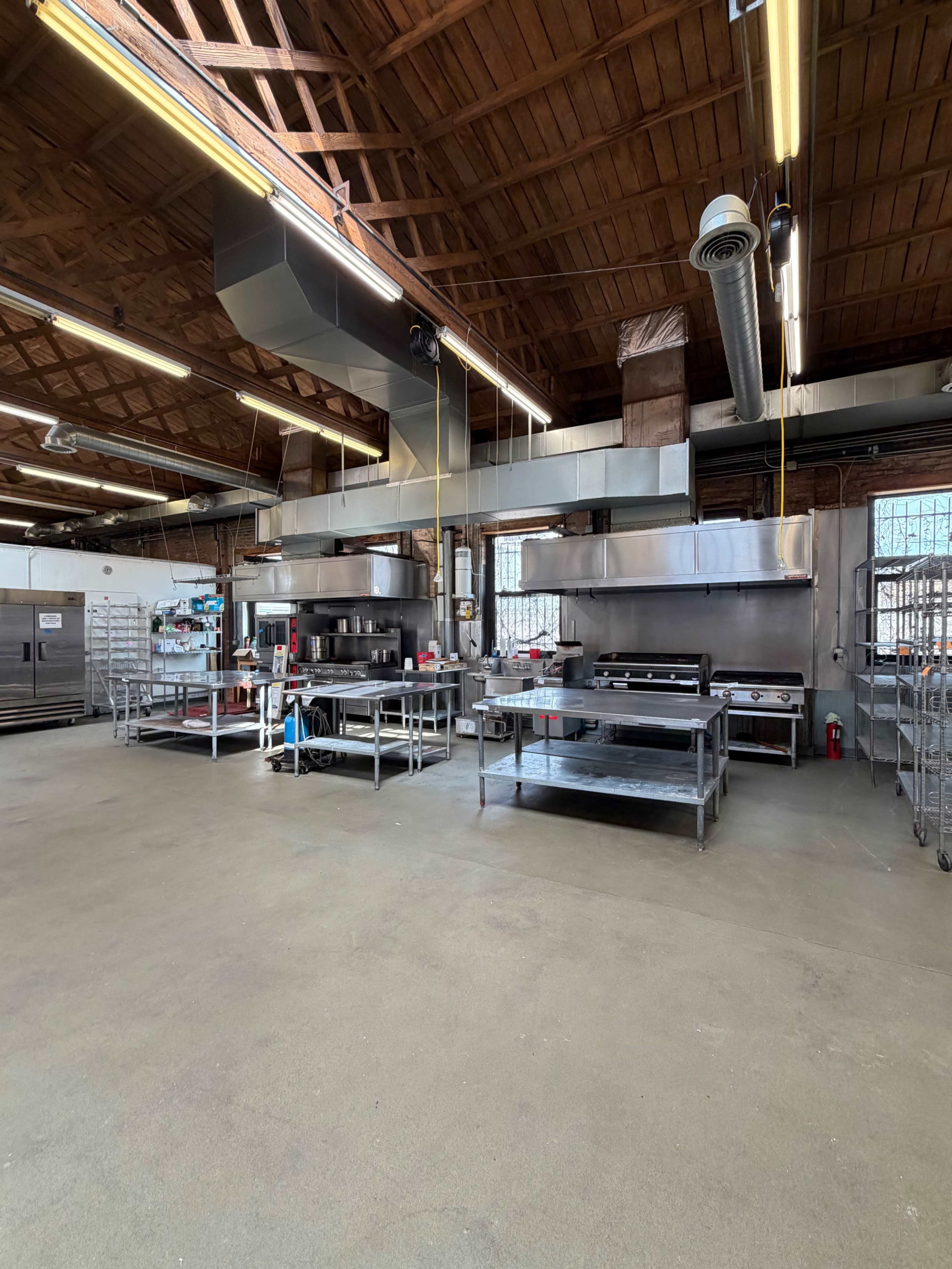 The image shows a spacious commercial kitchen with stainless steel appliances and worktables, featuring exposed wooden beams and overhead ventilation.