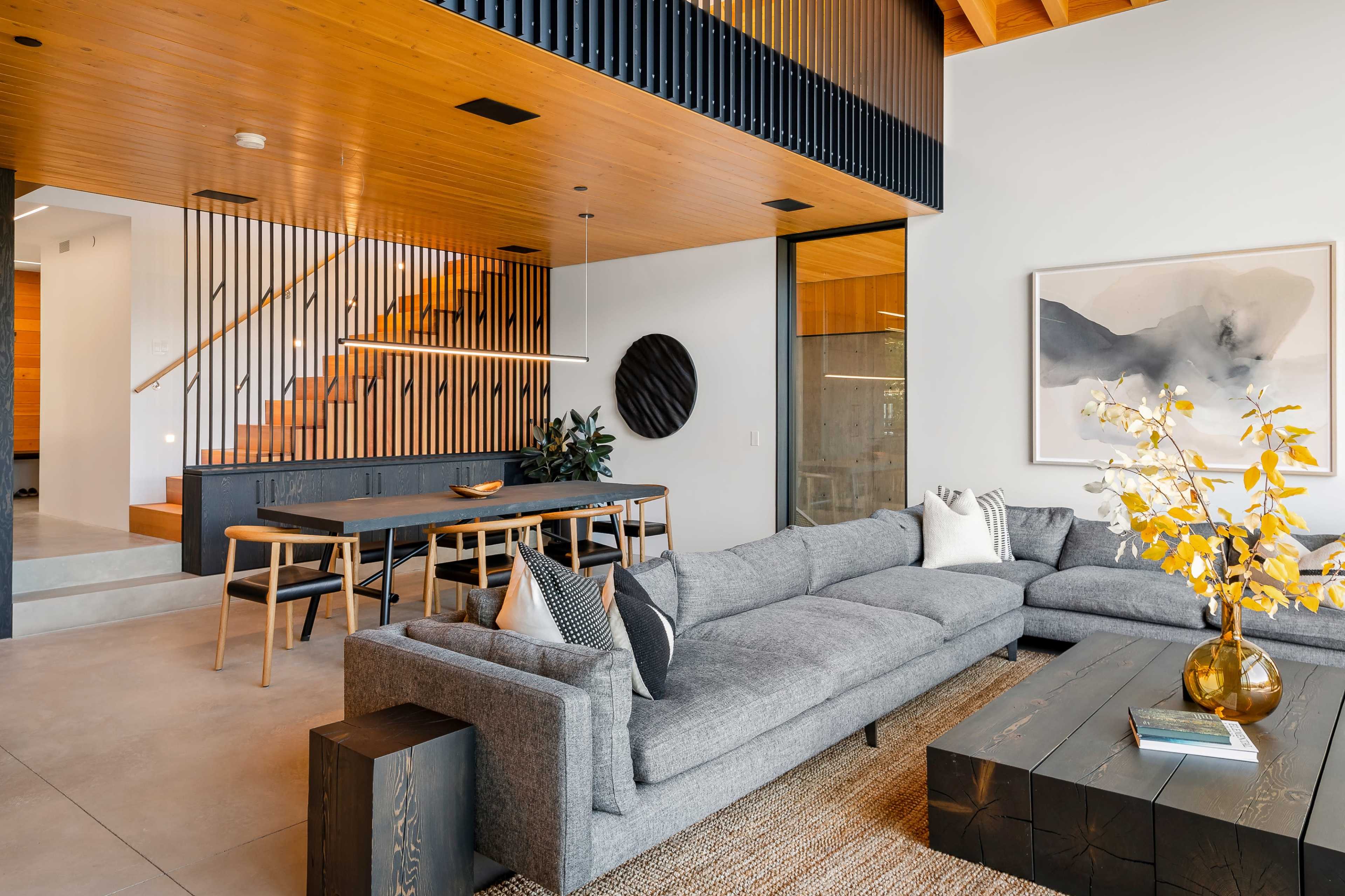 The image shows a modern living room with a gray sectional sofa, a wooden dining table, and an open staircase leading to an upper level.