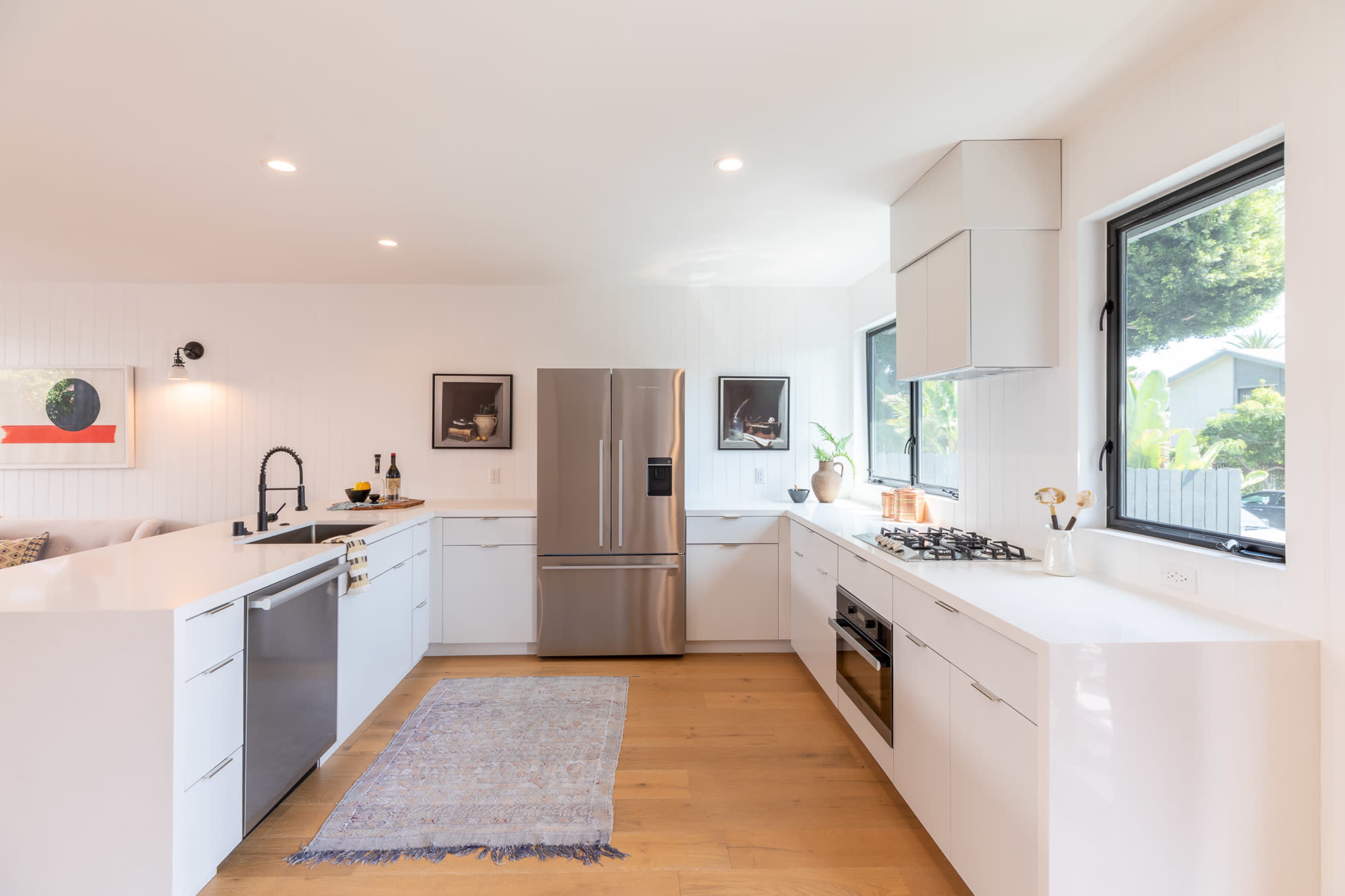 A modern kitchen features a stainless steel refrigerator, sleek white cabinetry, and a gas stove, illuminated by natural light from large windows.
