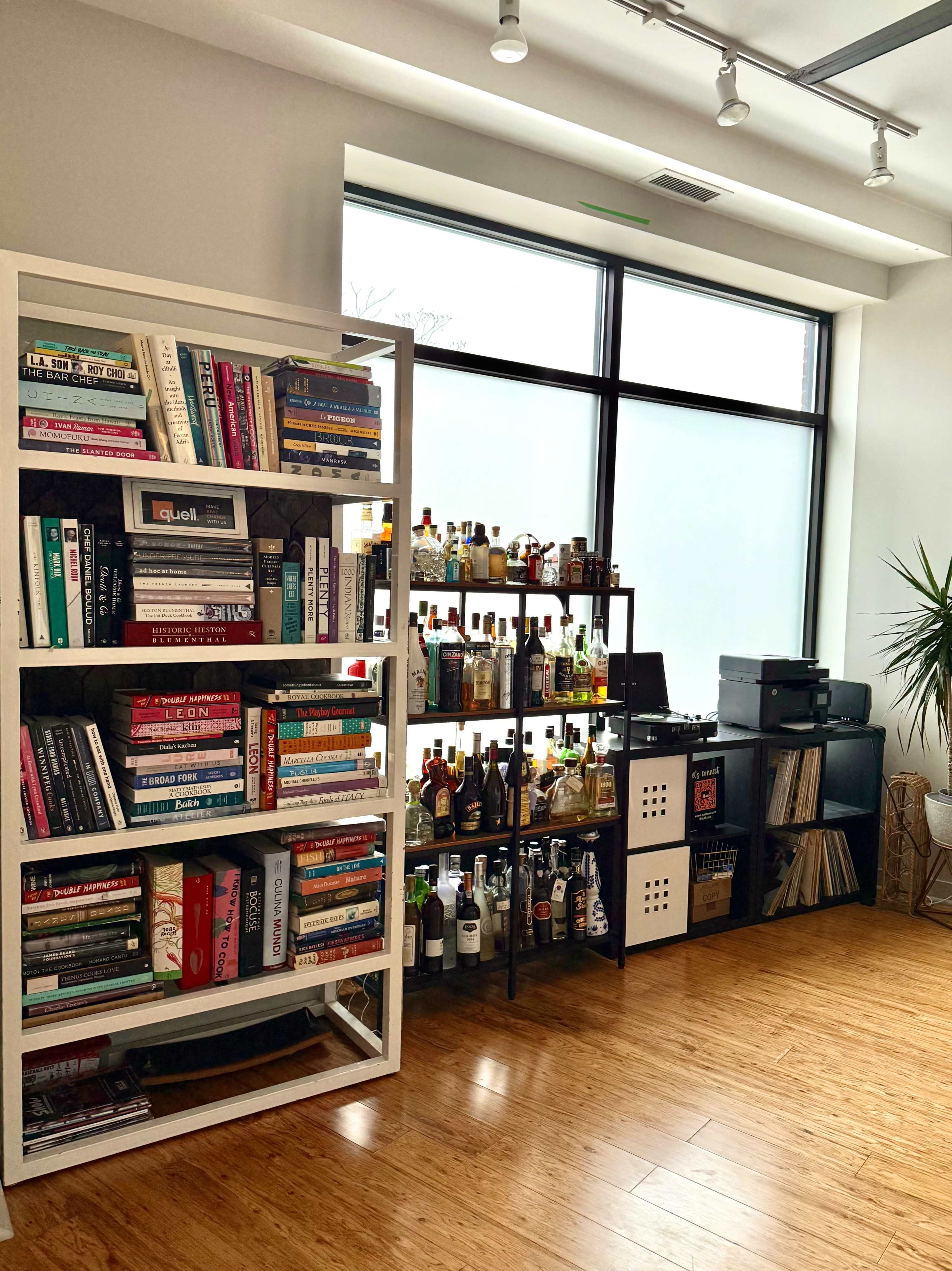 The image shows a well-organized room with a large bookshelf filled with various books on one side and a shelf displaying numerous bottles of liquor, along with a printer and vinyl records on the other side.