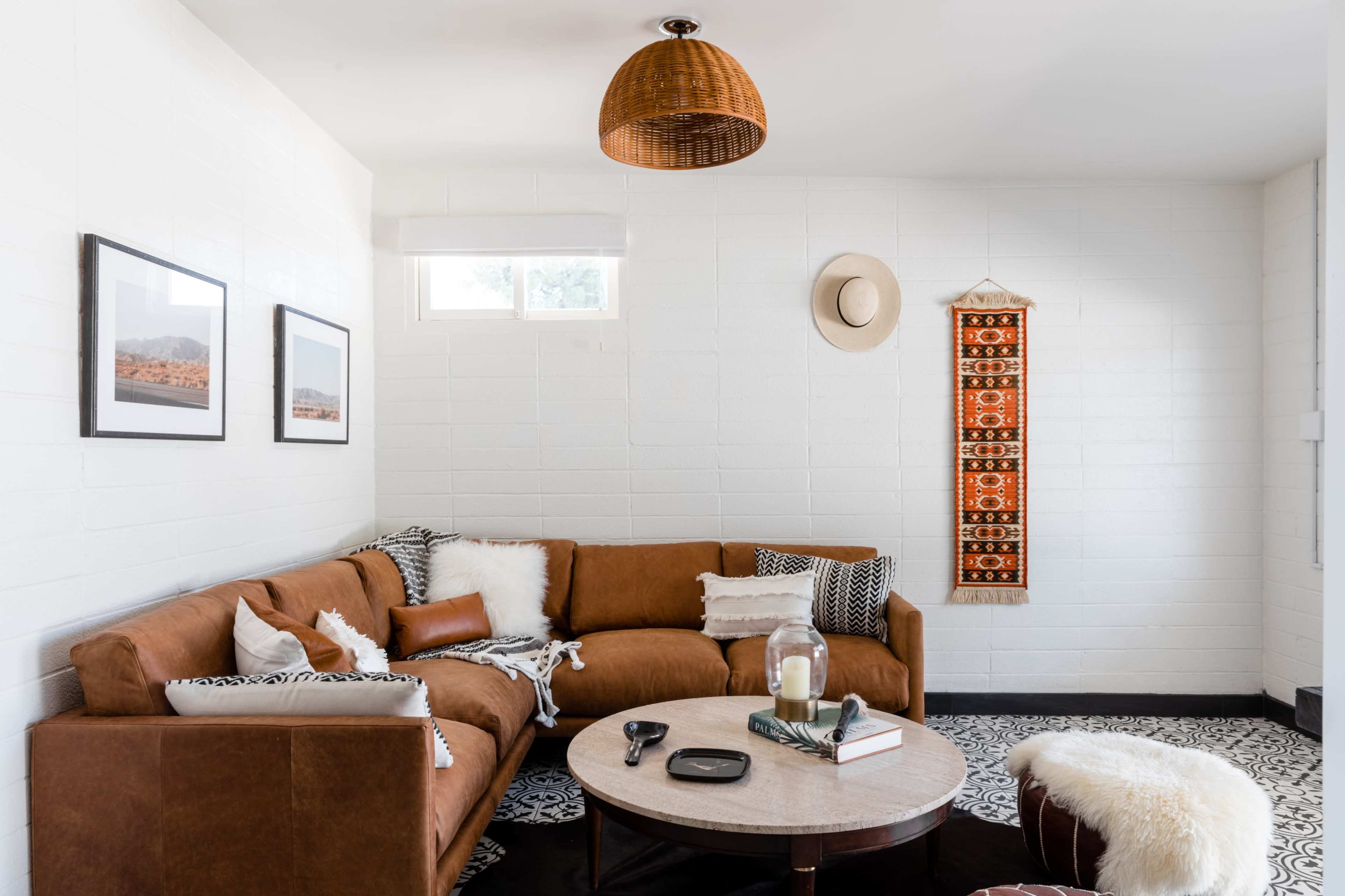 A cozy living room features a brown leather sectional sofa, a round coffee table, framed wall art, and decorative accents, with a woven pendant light overhead.