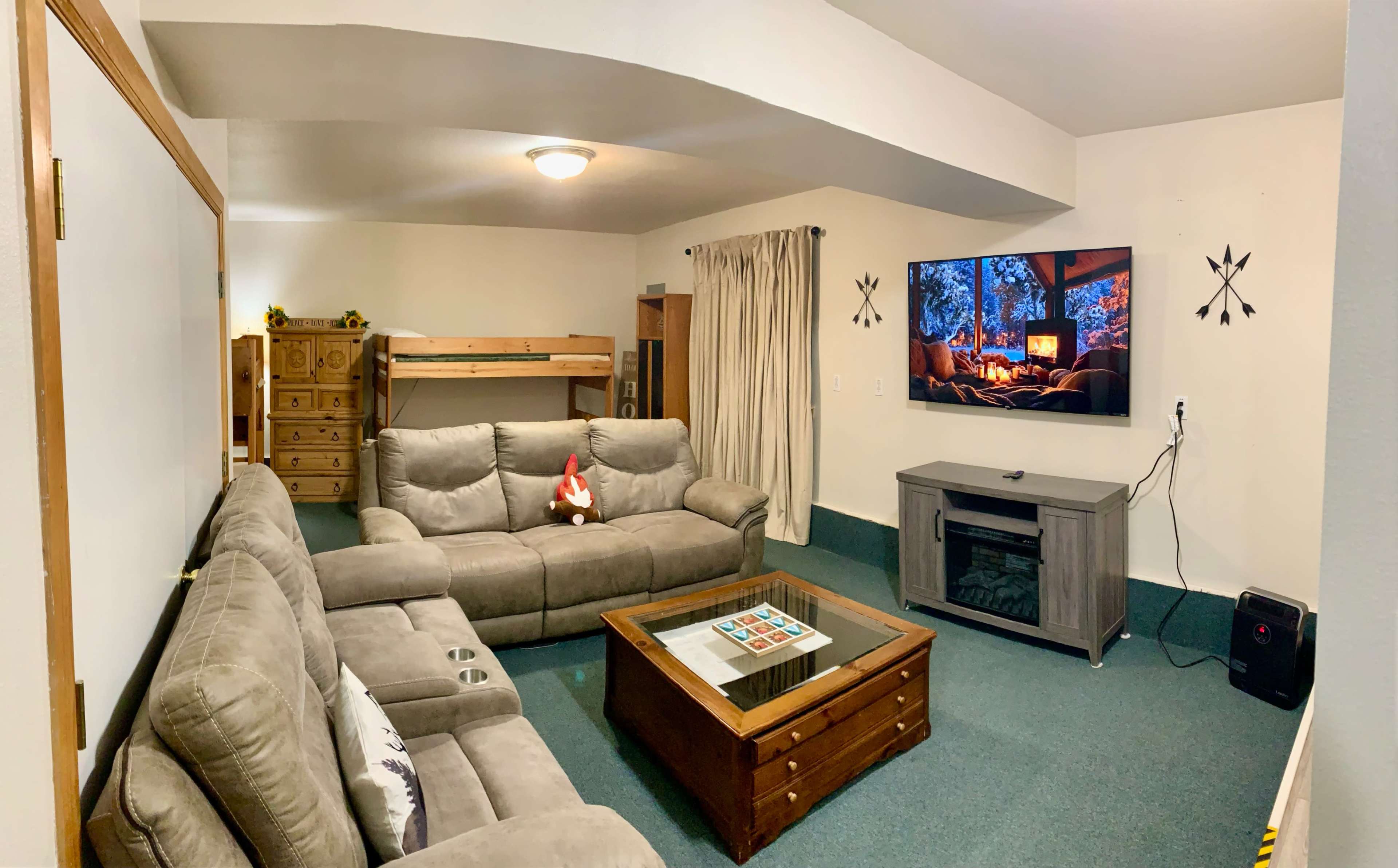 The image shows a cozy living room with a gray sectional sofa, a wooden coffee table, a television mounted on the wall, and a bunk bed in the background.