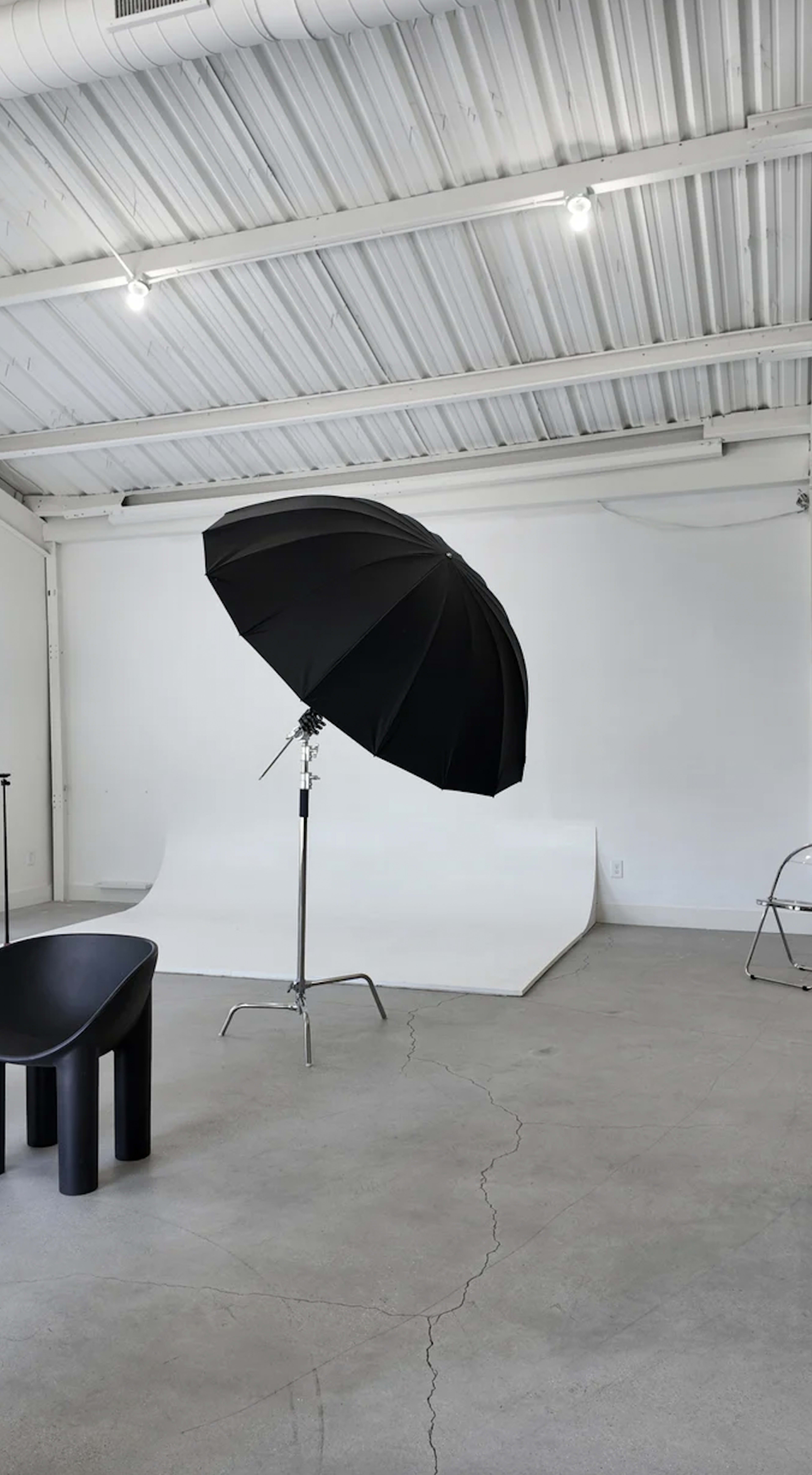 The image shows a spacious photography studio featuring a black umbrella softbox, a black chair, and a white backdrop on the floor.