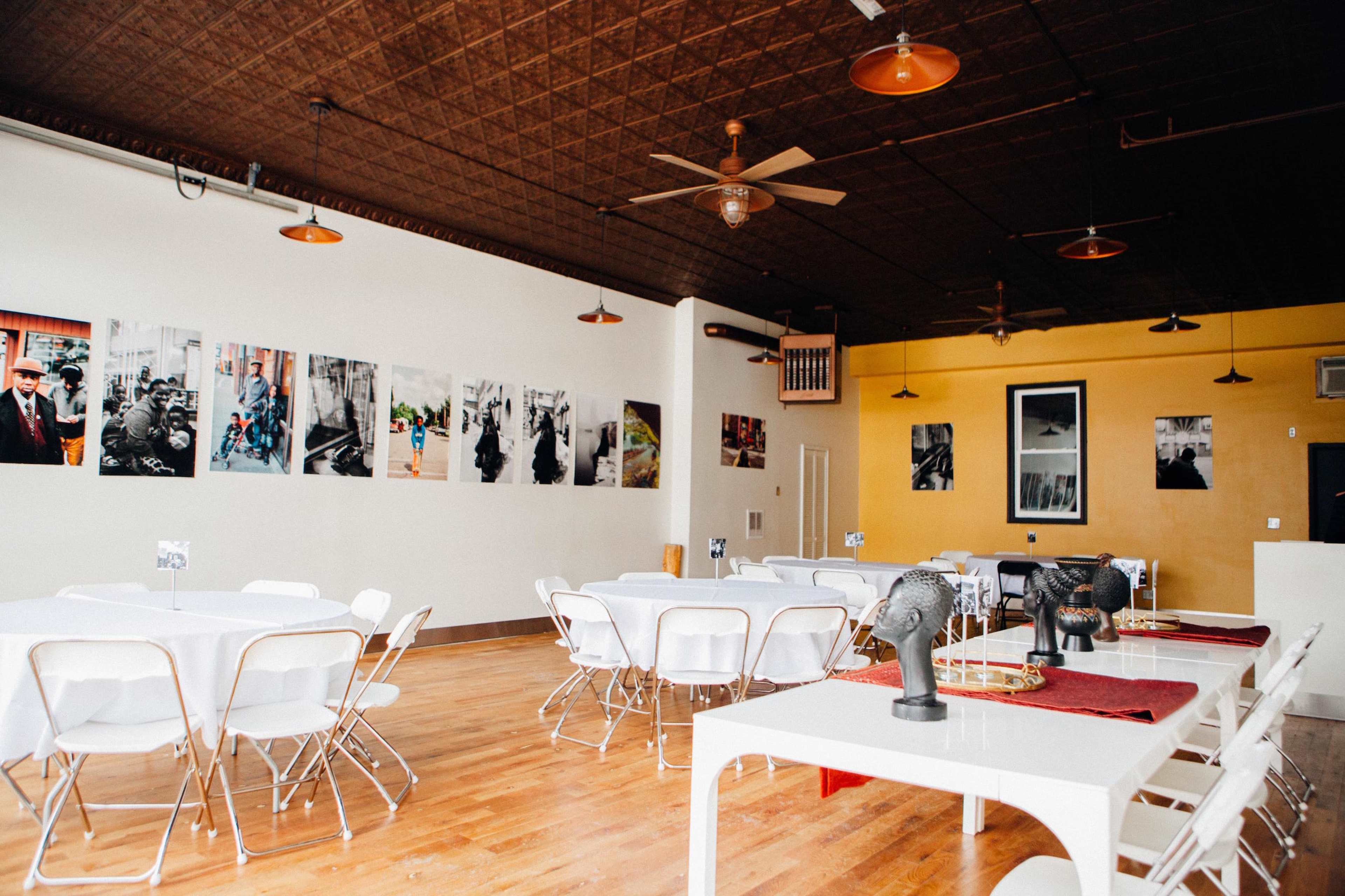A spacious, well-lit restaurant interior with several tables set up, wall art featuring black and white photographs, and ceiling fans.