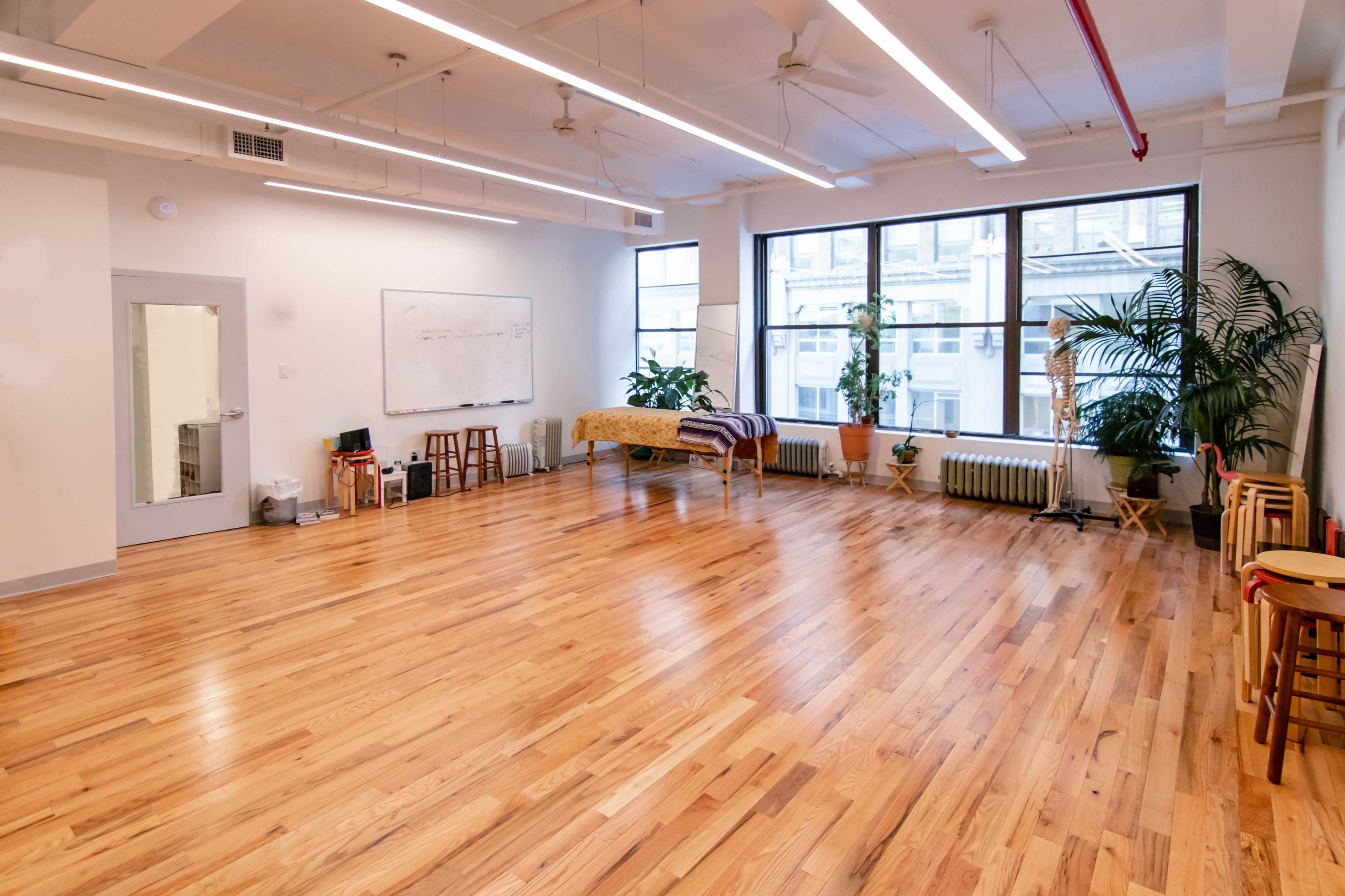 A spacious studio with wooden flooring, large windows, several chairs, and a whiteboard on the wall.