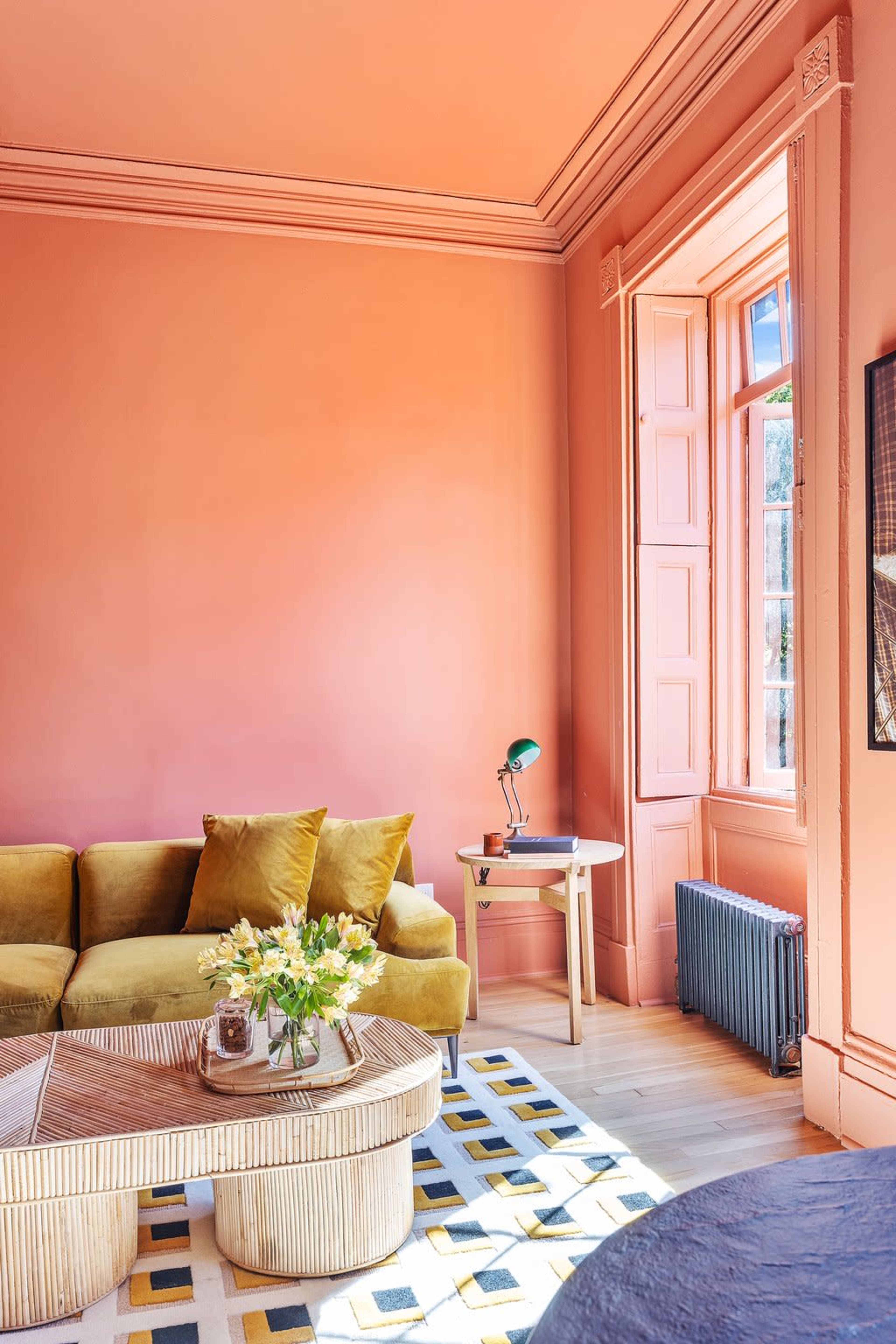 The image shows a living room with a peach-colored wall, a green sofa, a round coffee table, and a window with pink frames allowing natural light to enter.