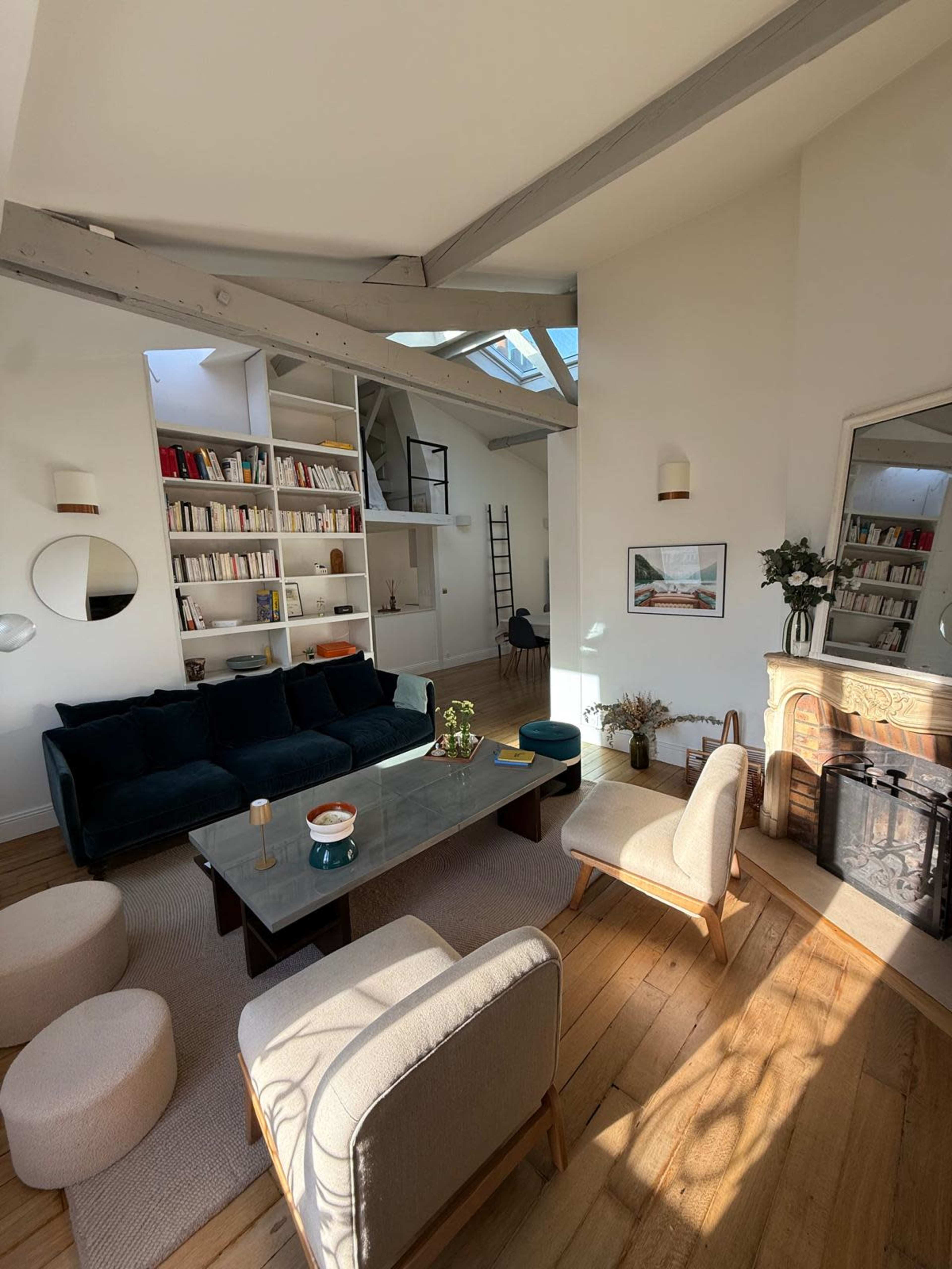 The image shows a modern living room with a black sofa, a coffee table, light-colored armchairs, a bookshelf filled with books, and a fireplace, all under a bright skylight.
