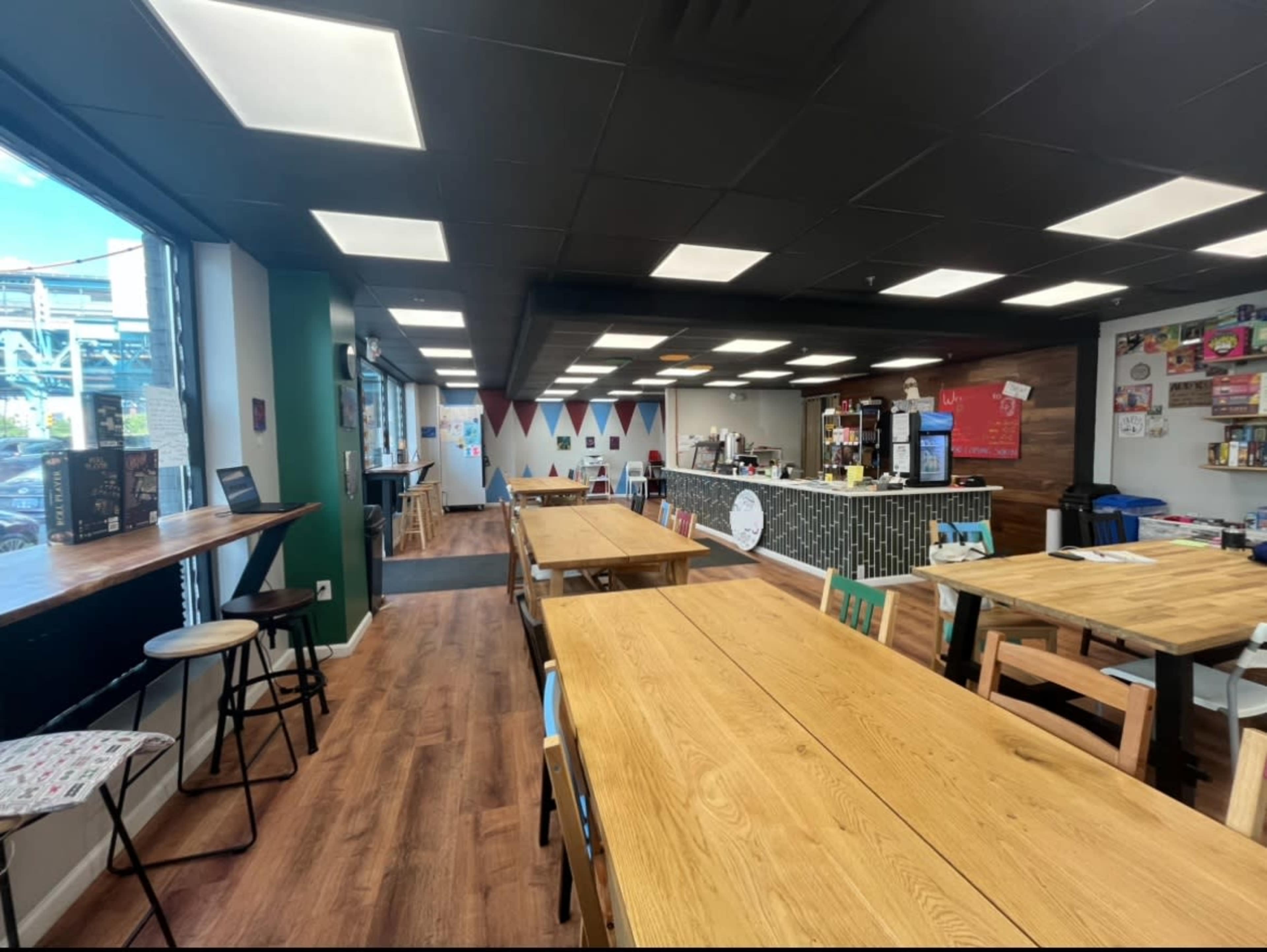 The image shows a spacious cafe with multiple wooden tables, chairs, a service counter, and decorative banners hanging from the ceiling.