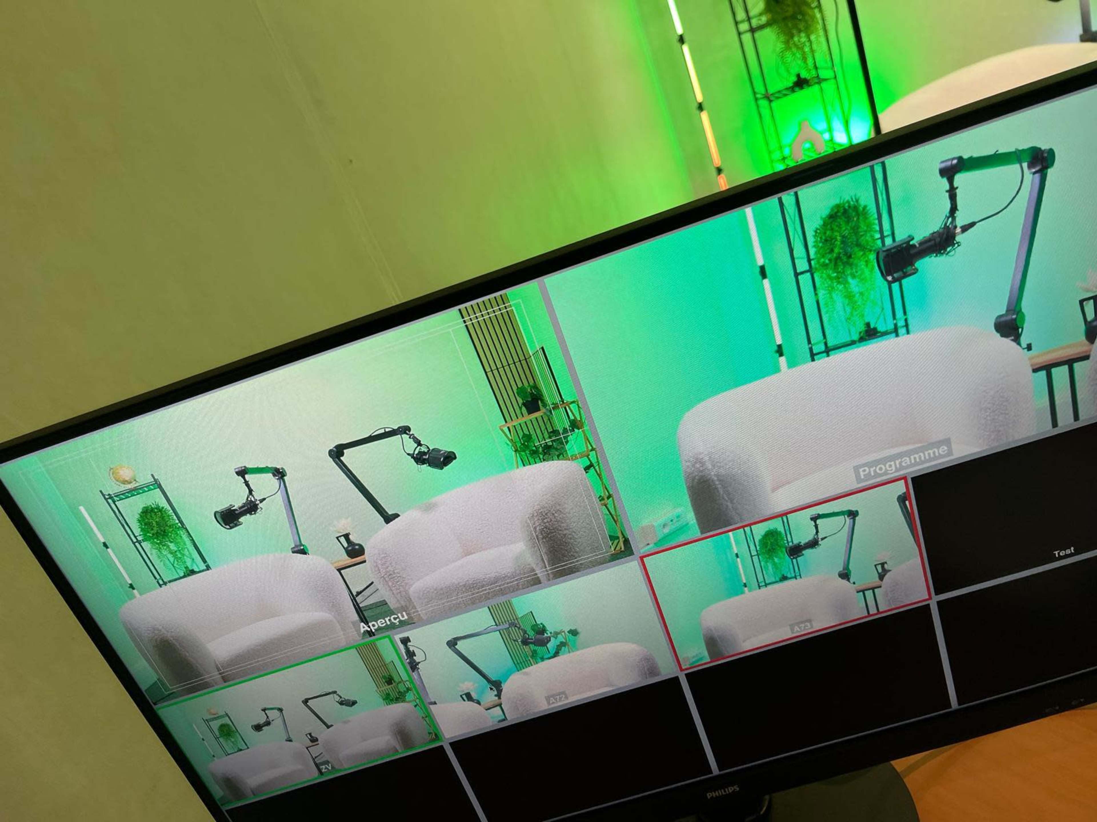 A monitor displays multiple camera feeds of a studio setup featuring green lighting and a white seating area.