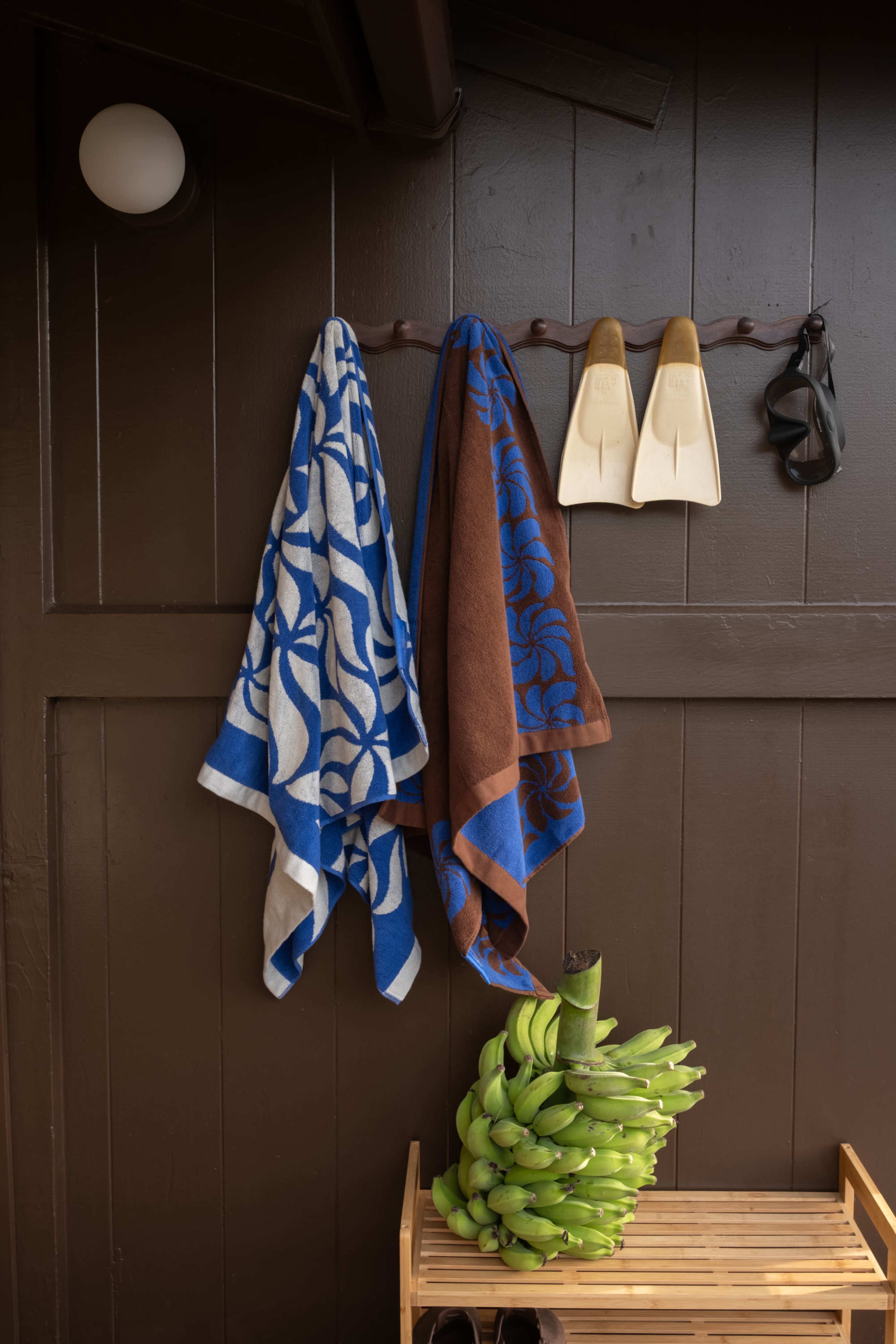 Two patterned towels hang on a wall hook next to a pair of wooden flip-flops, while a bunch of bananas sits on a wooden shelf below.