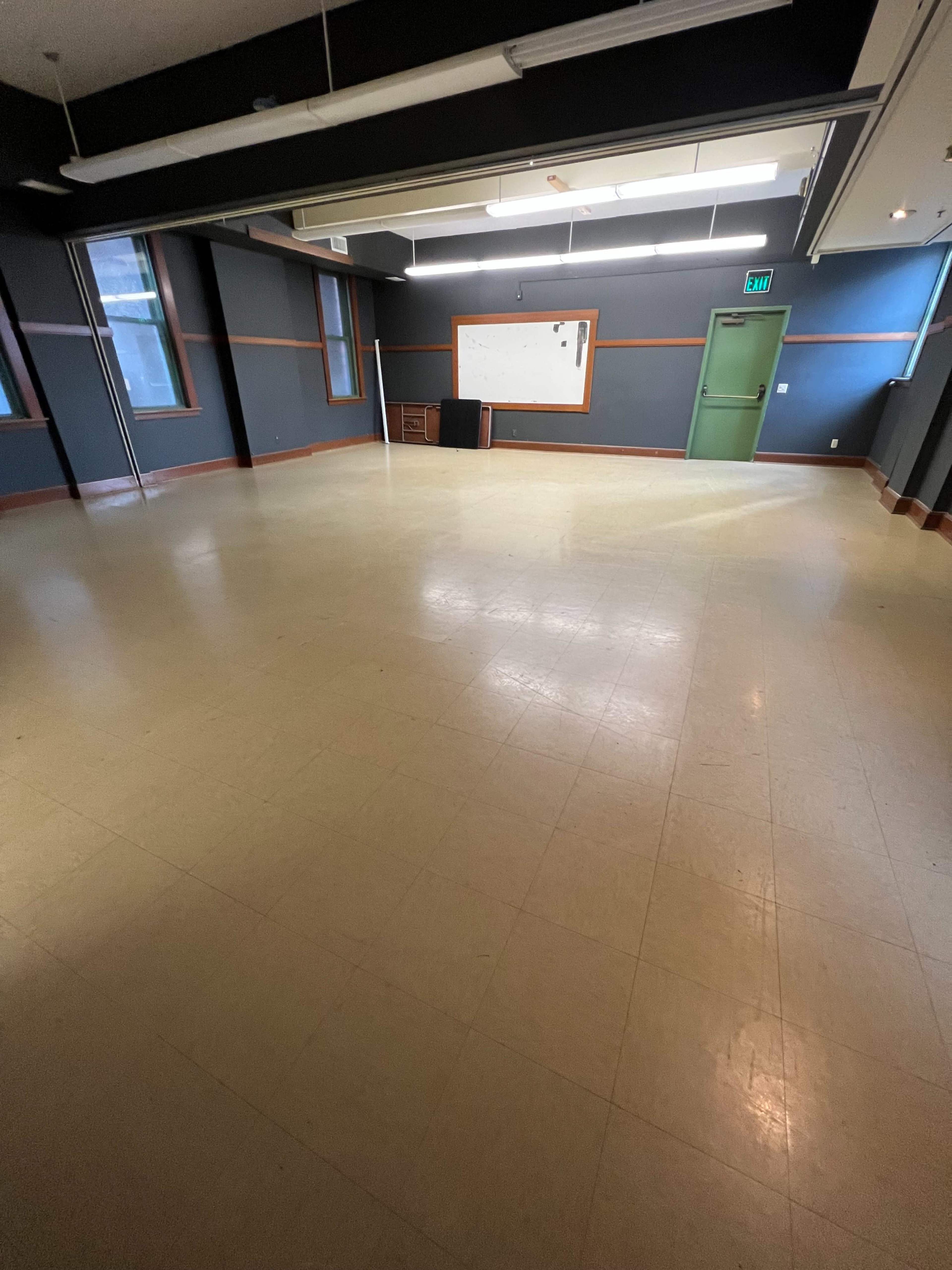 The image shows an empty classroom with a bare floor, a whiteboard on the wall, and a closed door.