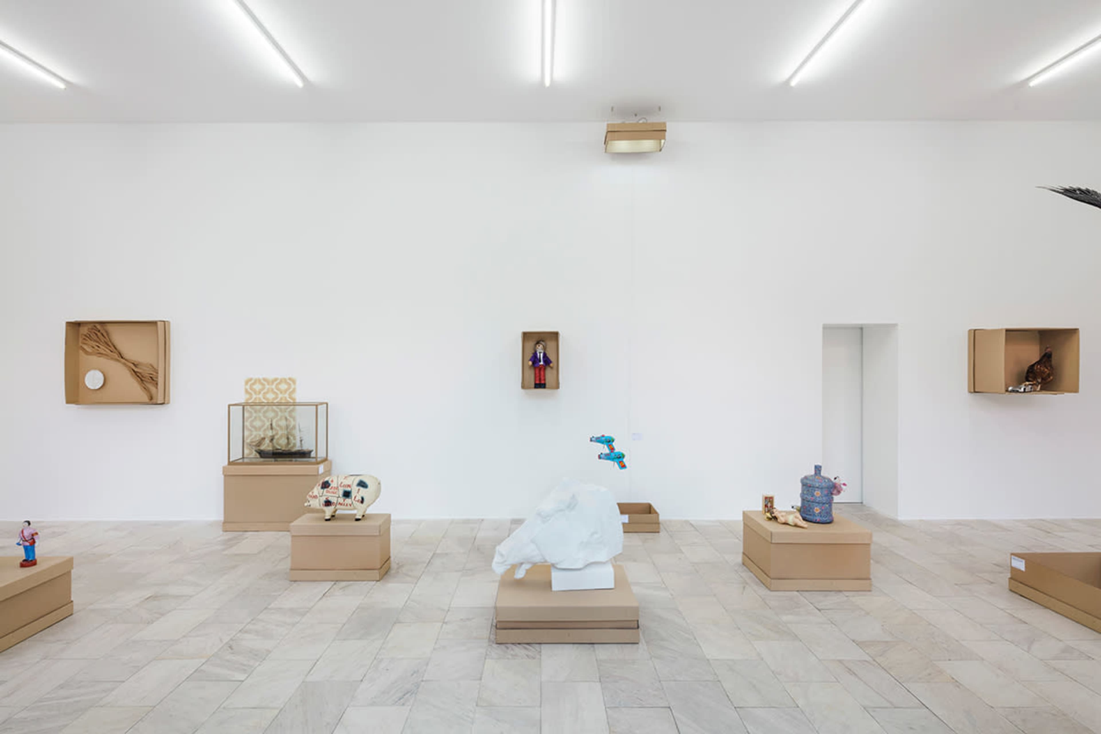 An art gallery with white walls and a light-colored tiled floor, featuring several display pedestals with diverse sculptures and artworks organized throughout the space.