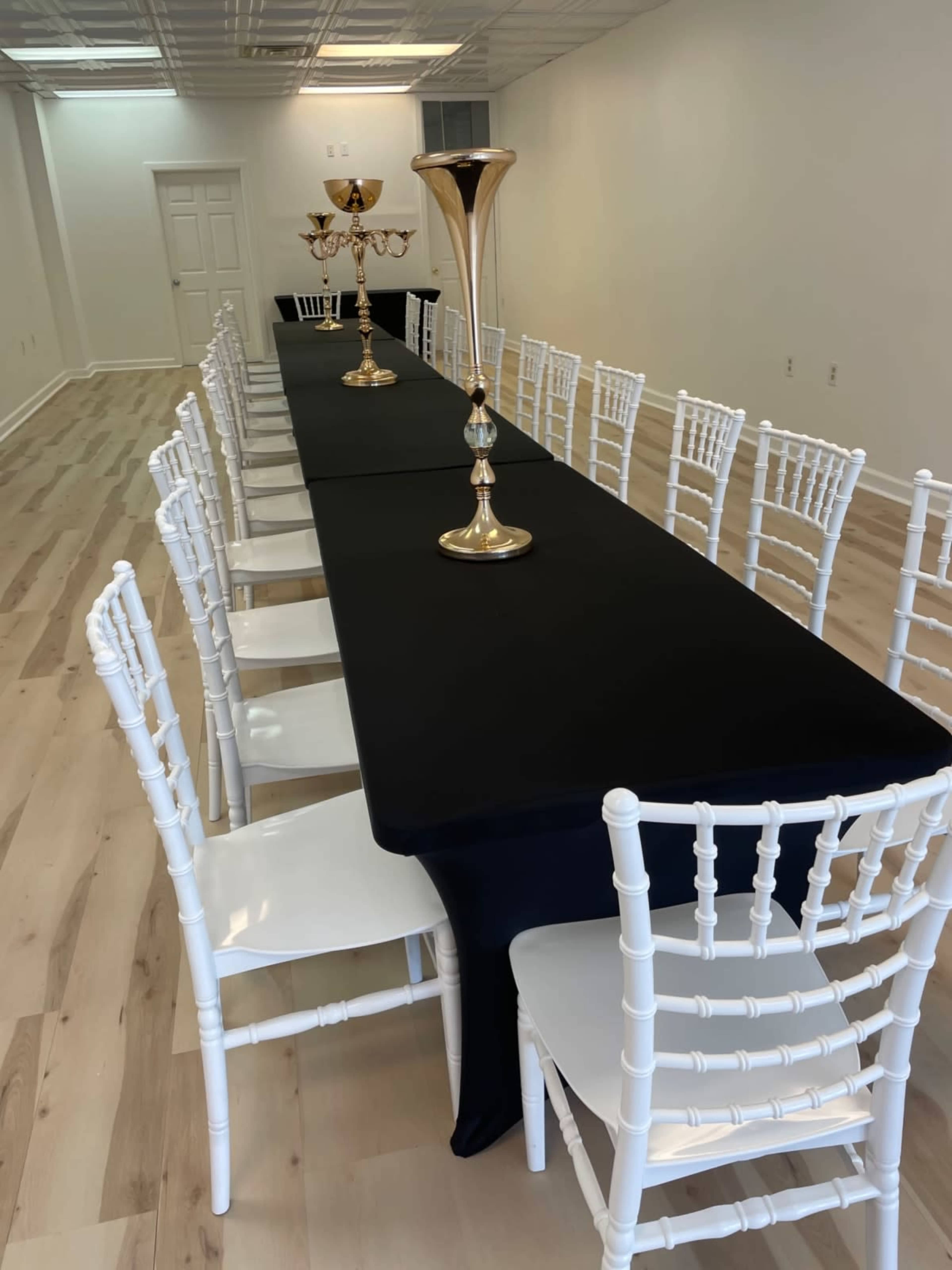 A long table covered with a black cloth is set up in a spacious, empty room, flanked by white chairs and adorned with gold candle holders.