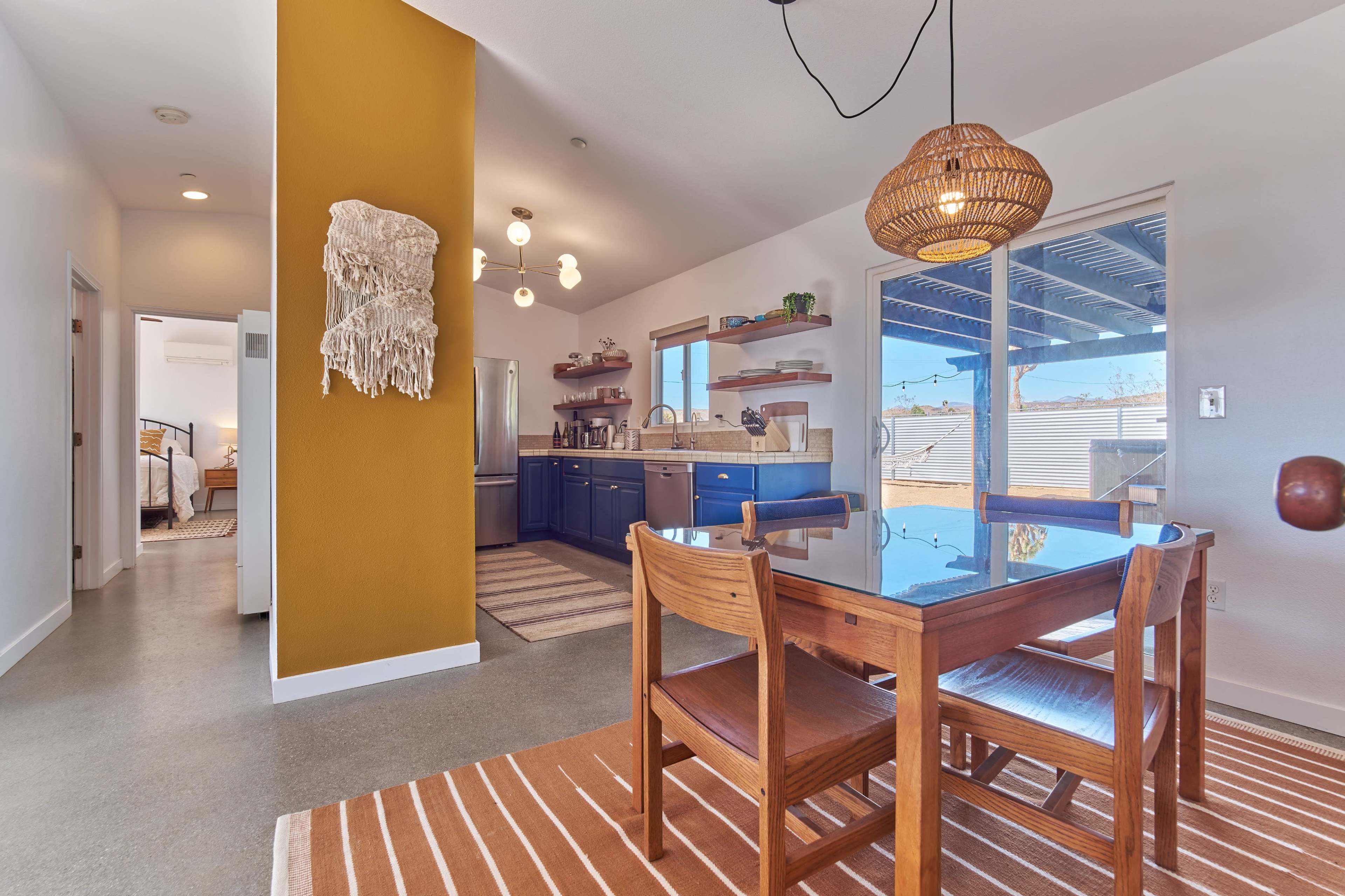A dining area features a wooden table and chairs, with a patterned rug, surrounded by a kitchen with blue cabinetry and an adjacent doorway leading to another room.