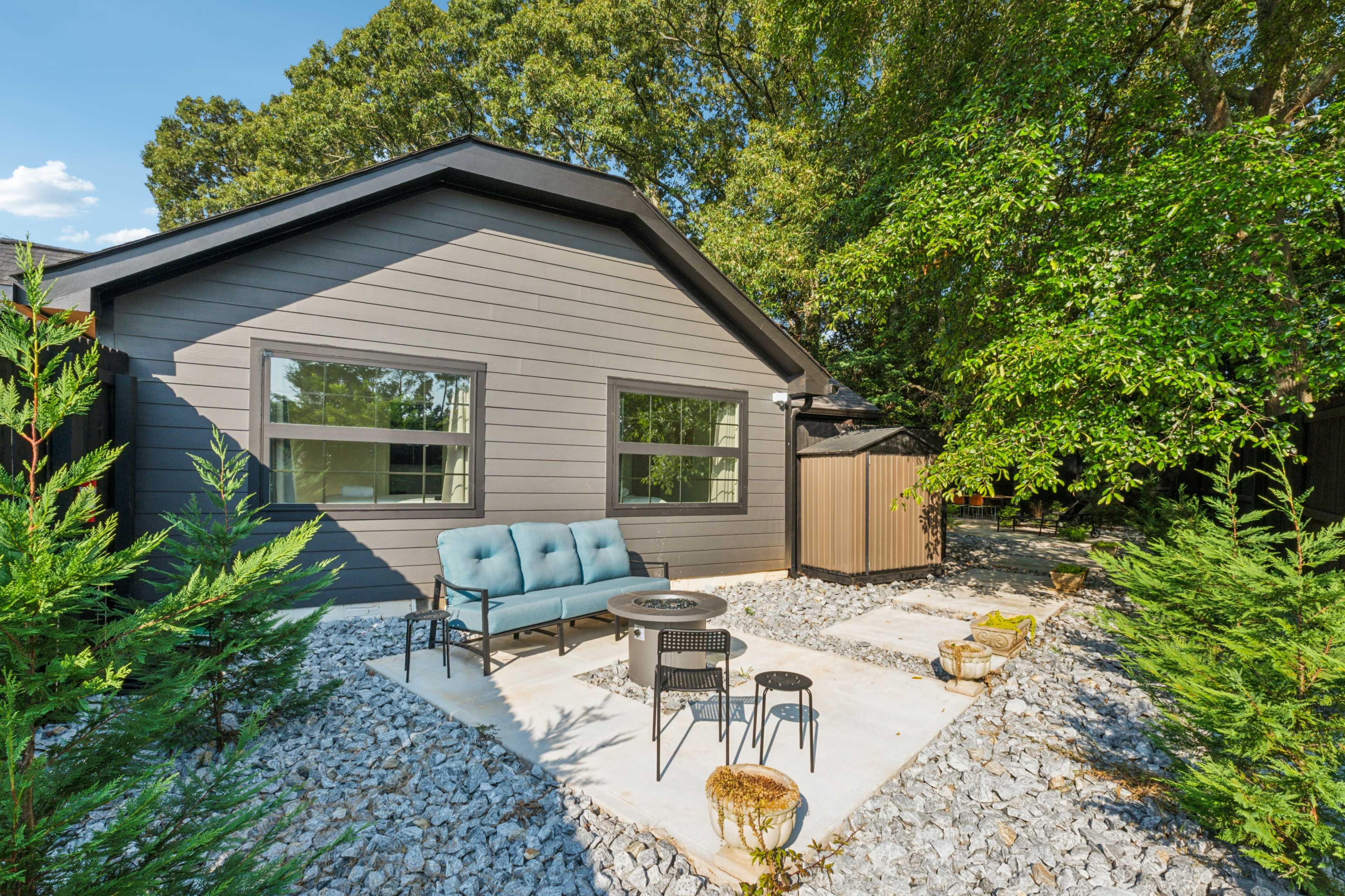 A modern, single-story house with a gray exterior features a patio area with seating and a fire pit, surrounded by gravel and greenery.