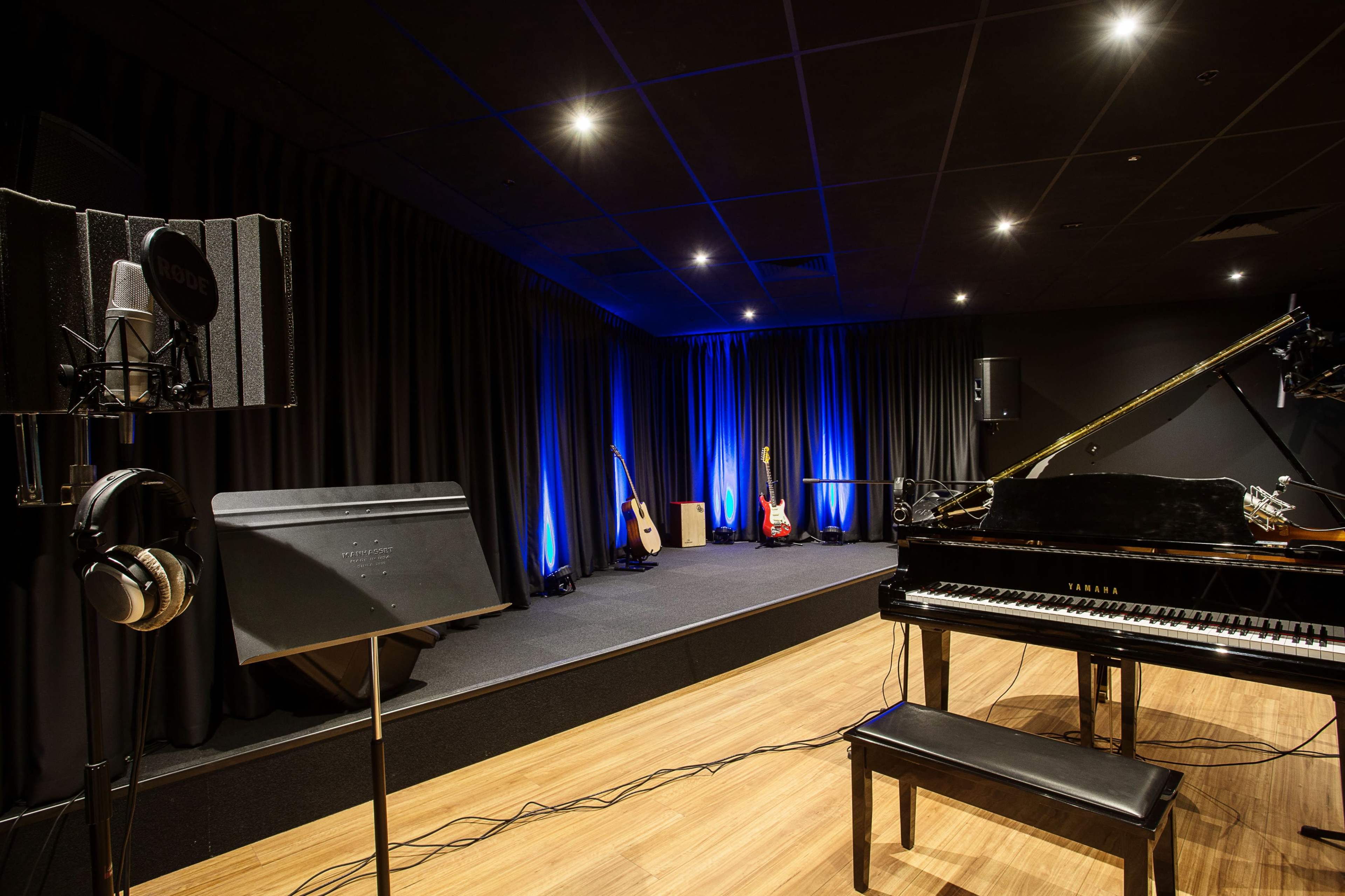 The image shows a music studio featuring a grand piano, microphones, audio equipment, and illuminated blue lights against a black curtain backdrop.