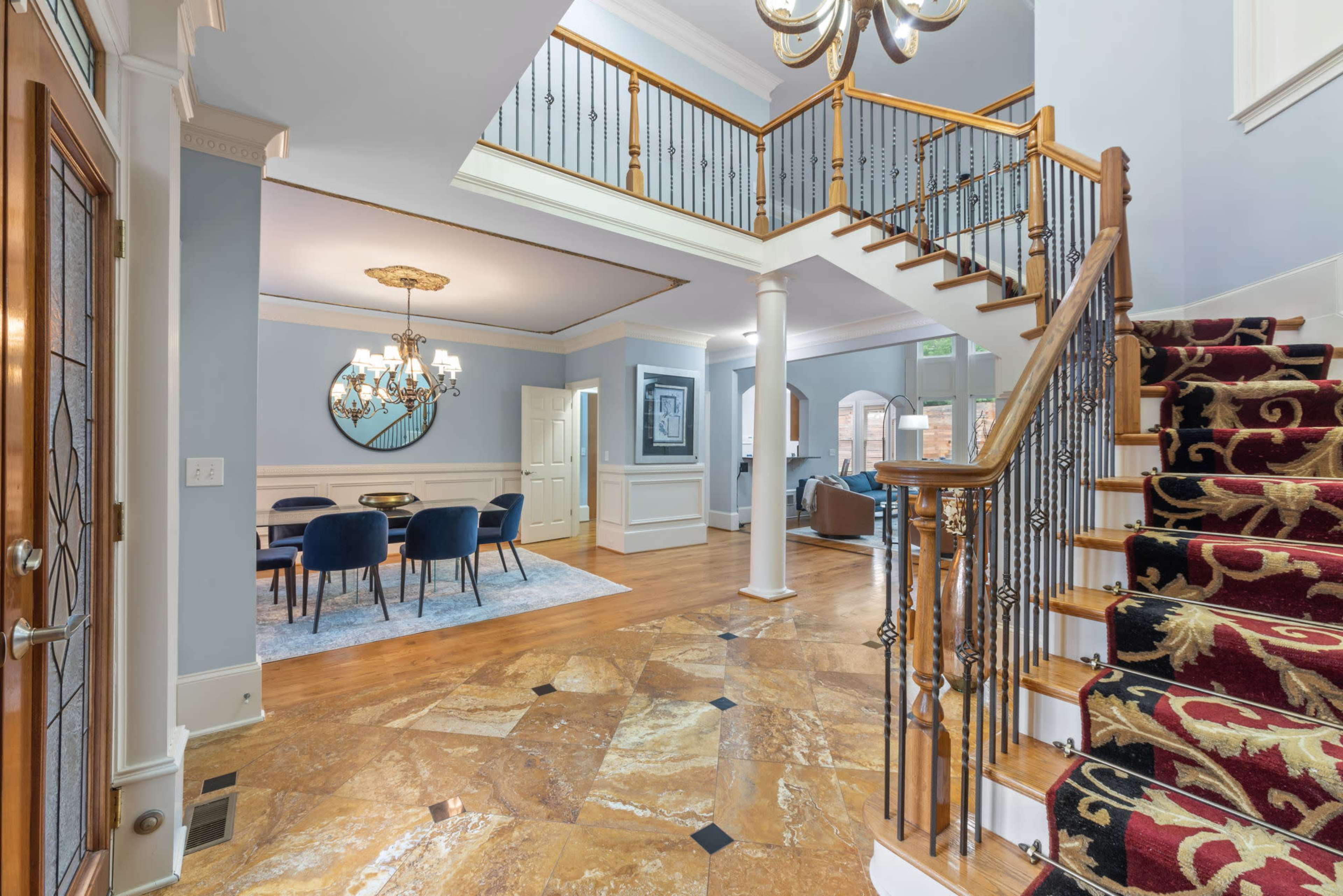 The image shows a spacious foyer with a winding staircase, hardwood flooring, and a chandelier, leading to a dining area in the background.