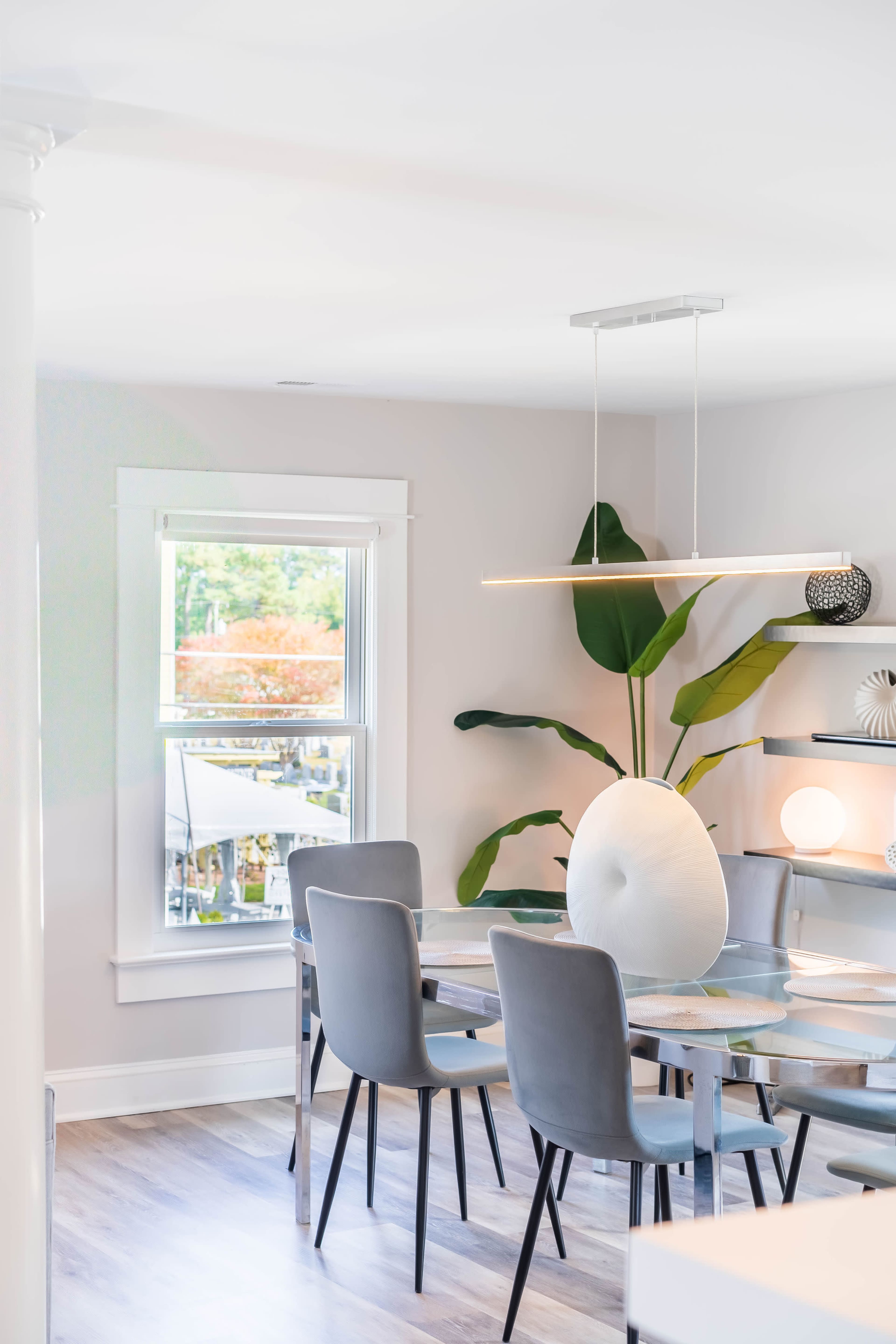 A modern dining area features a glass table surrounded by four gray chairs, with a large decorative vase and a tall plant nearby.