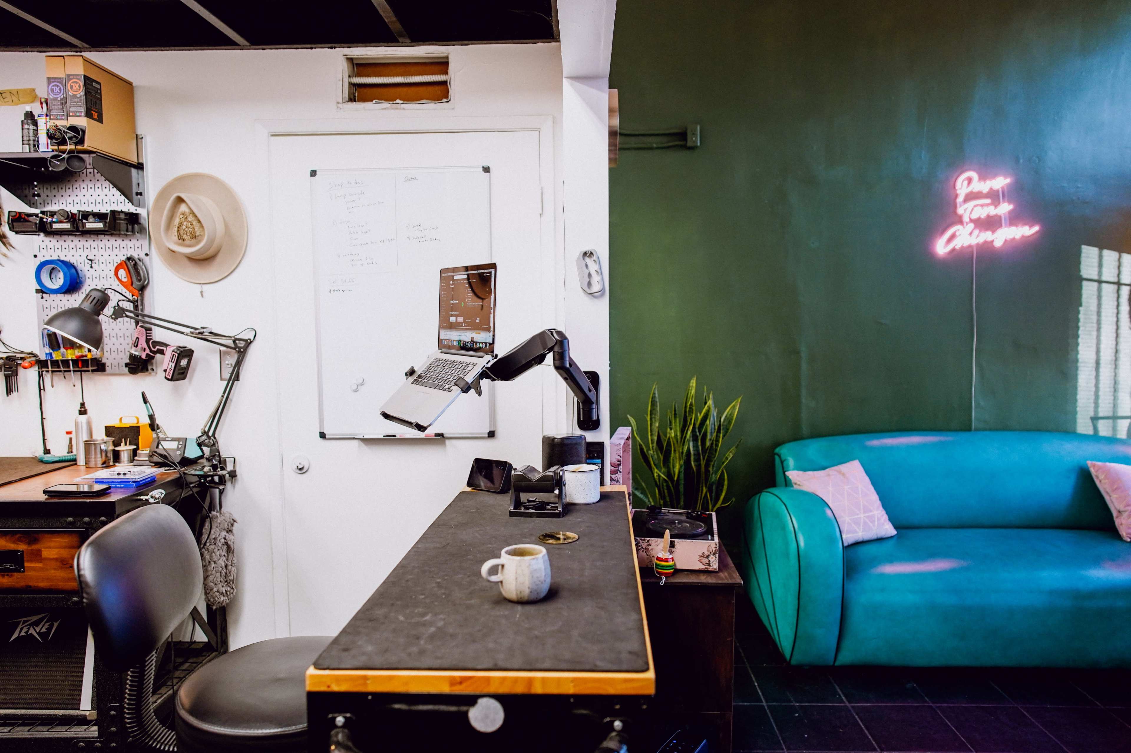 The image shows a split scene of a workspace on the left with a desk, chair, and tools, and a lounge area on the right featuring a teal sofa and a neon sign.