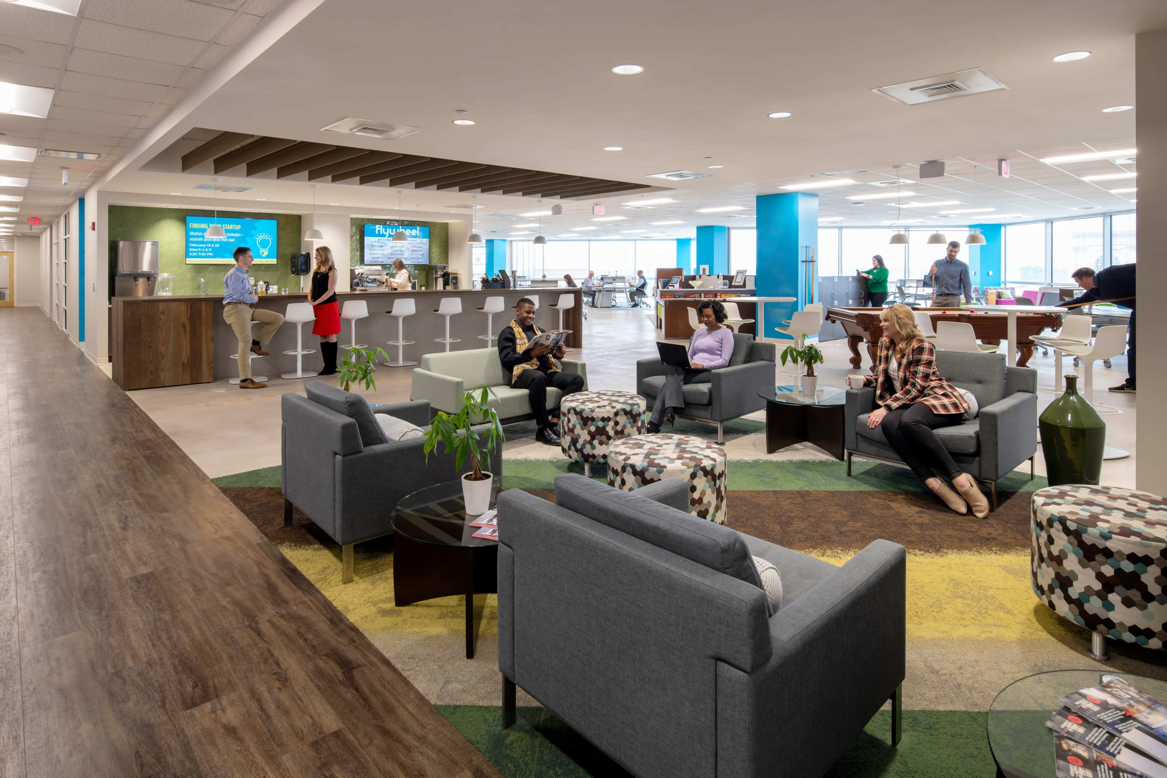 A modern office space with people engaged in various activities, featuring a lounge area with seating, a kitchen bar, and workstations in the background.