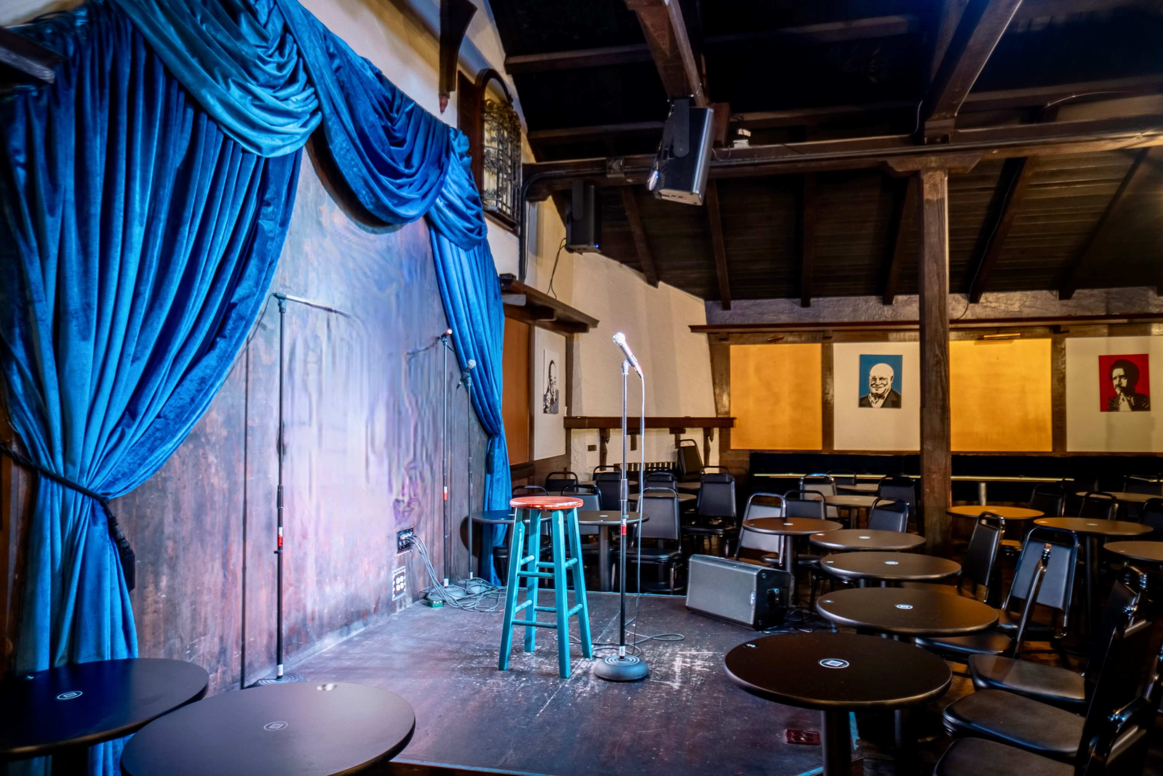 A comedy stage with a microphone on a stool is set in a dimly lit room featuring empty tables and artwork on the walls.