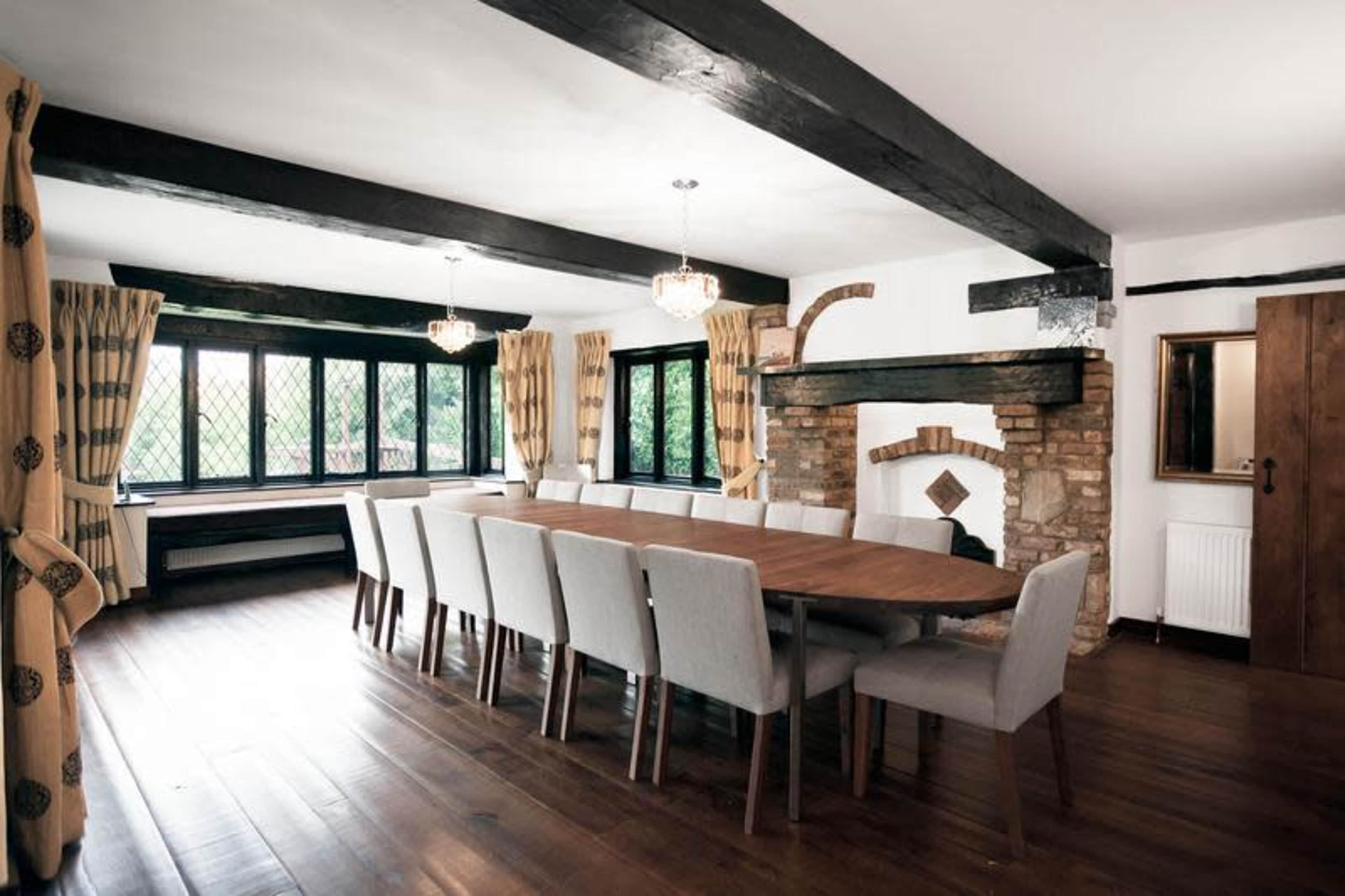 A spacious dining room features a long wooden table surrounded by upholstered chairs, large windows with rich curtains, and exposed wooden beams on the ceiling.